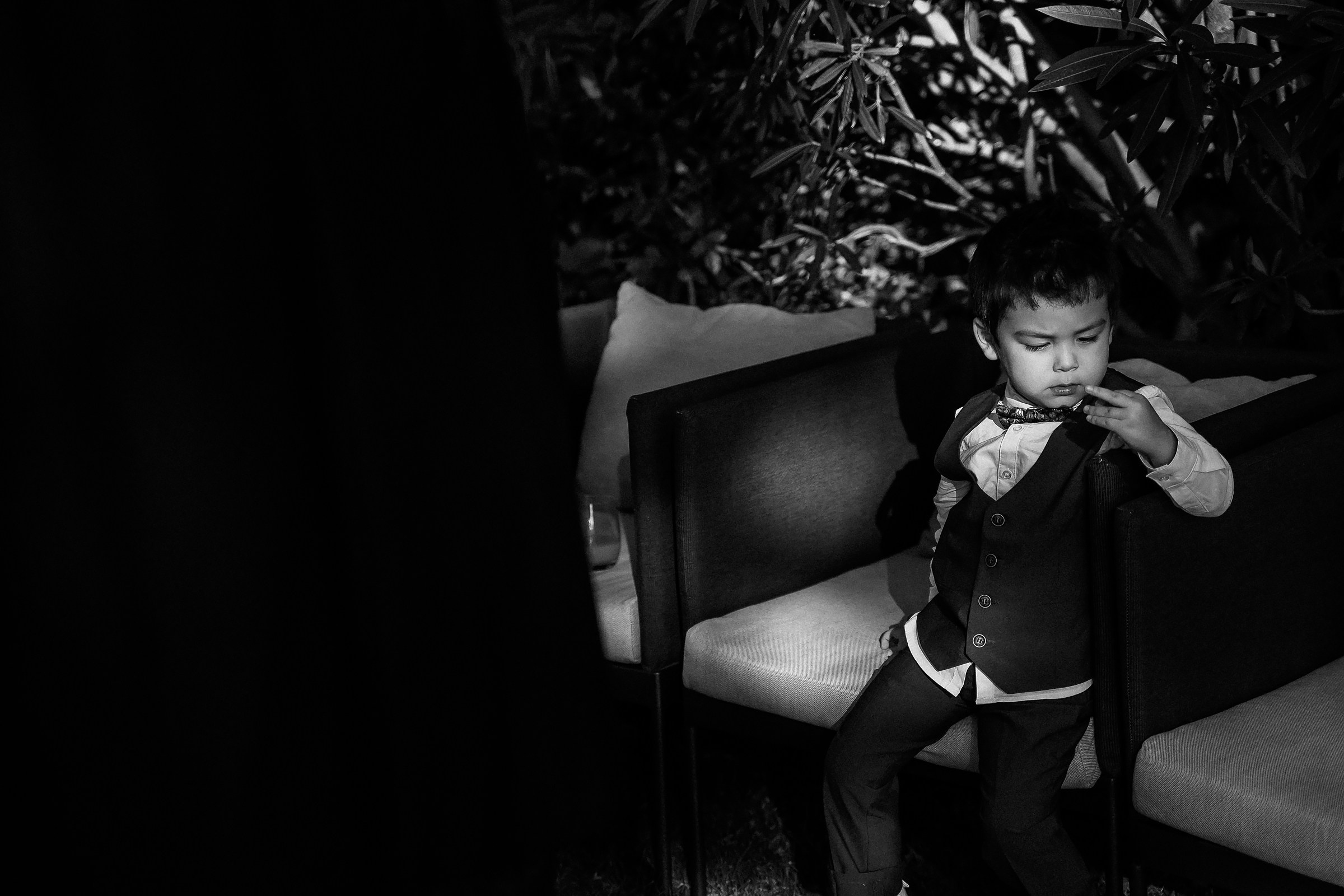 Young boy in formal attire sitting alone on a chair, looking thoughtful in a shaded garden setting.