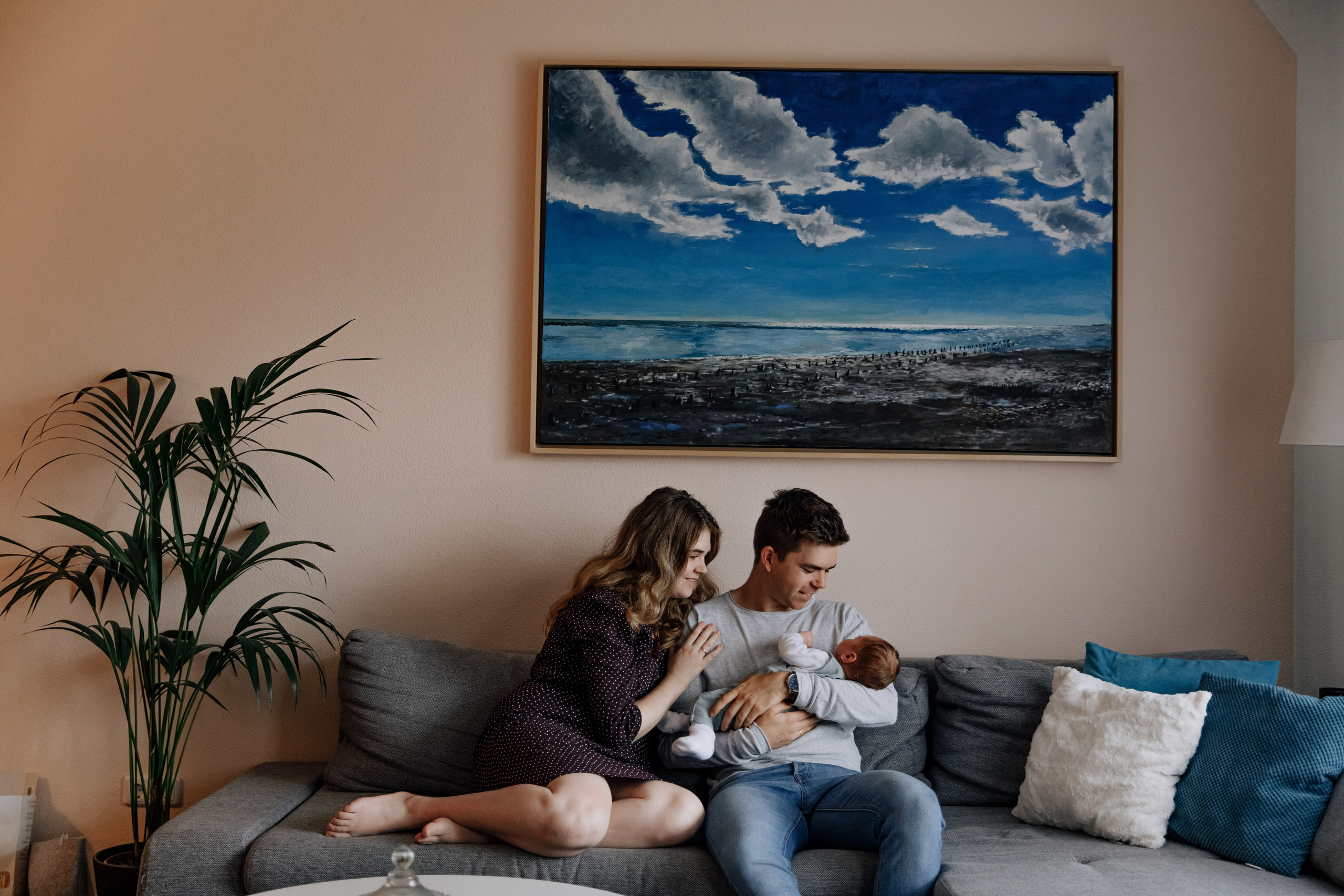 Eltern sitzen mit Baby auf dem Sofa zu Hause, im Hintergrund ein schönes Meeresbild an der Wand.
