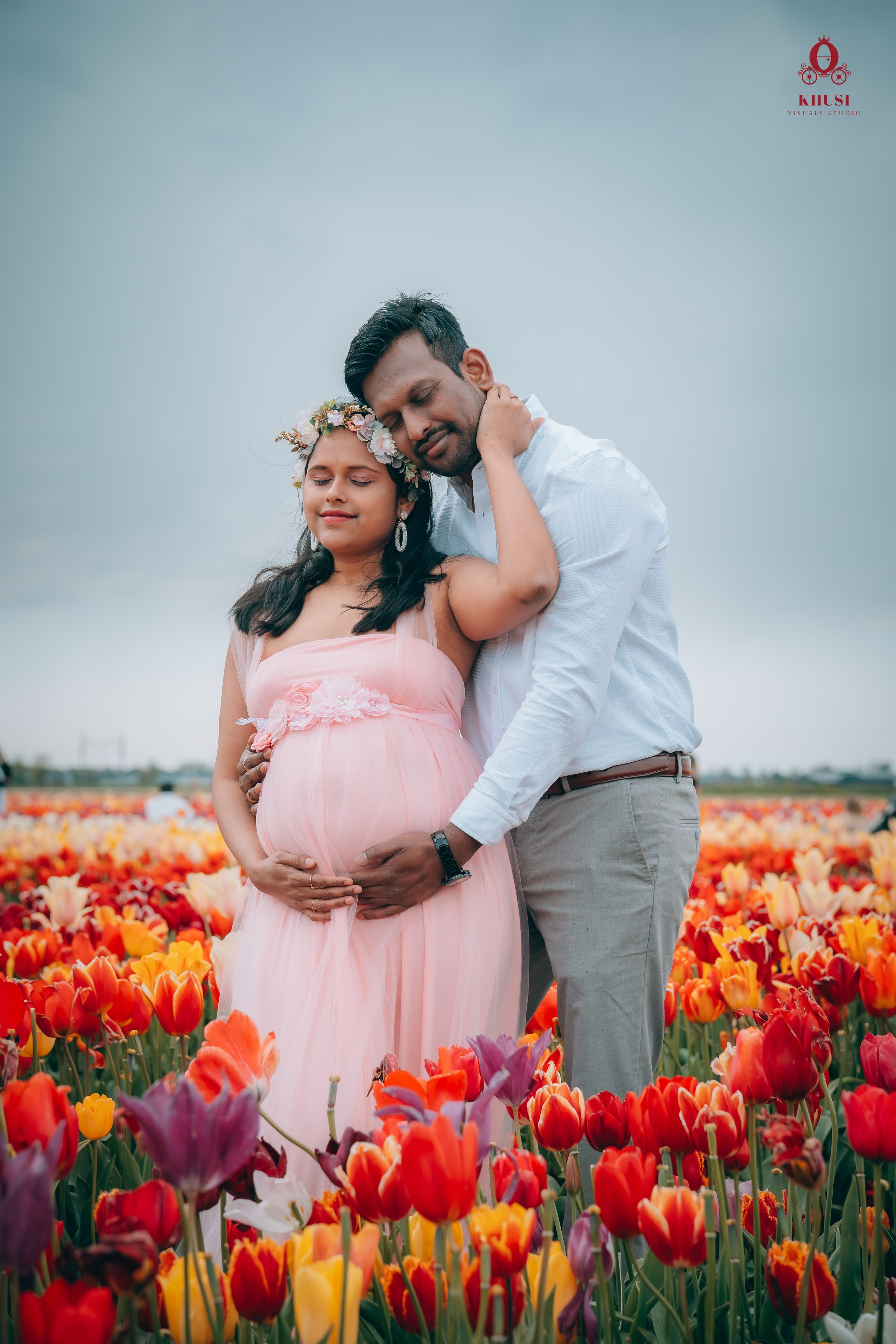A pregnant couple standing in a tulip flower field and hugging each other