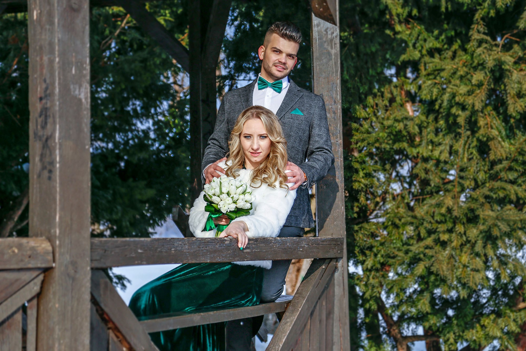 Wide shot of the bride and groom posing inside a rustic wooden gazebo surrounded by snowy trees during their photo shoot.