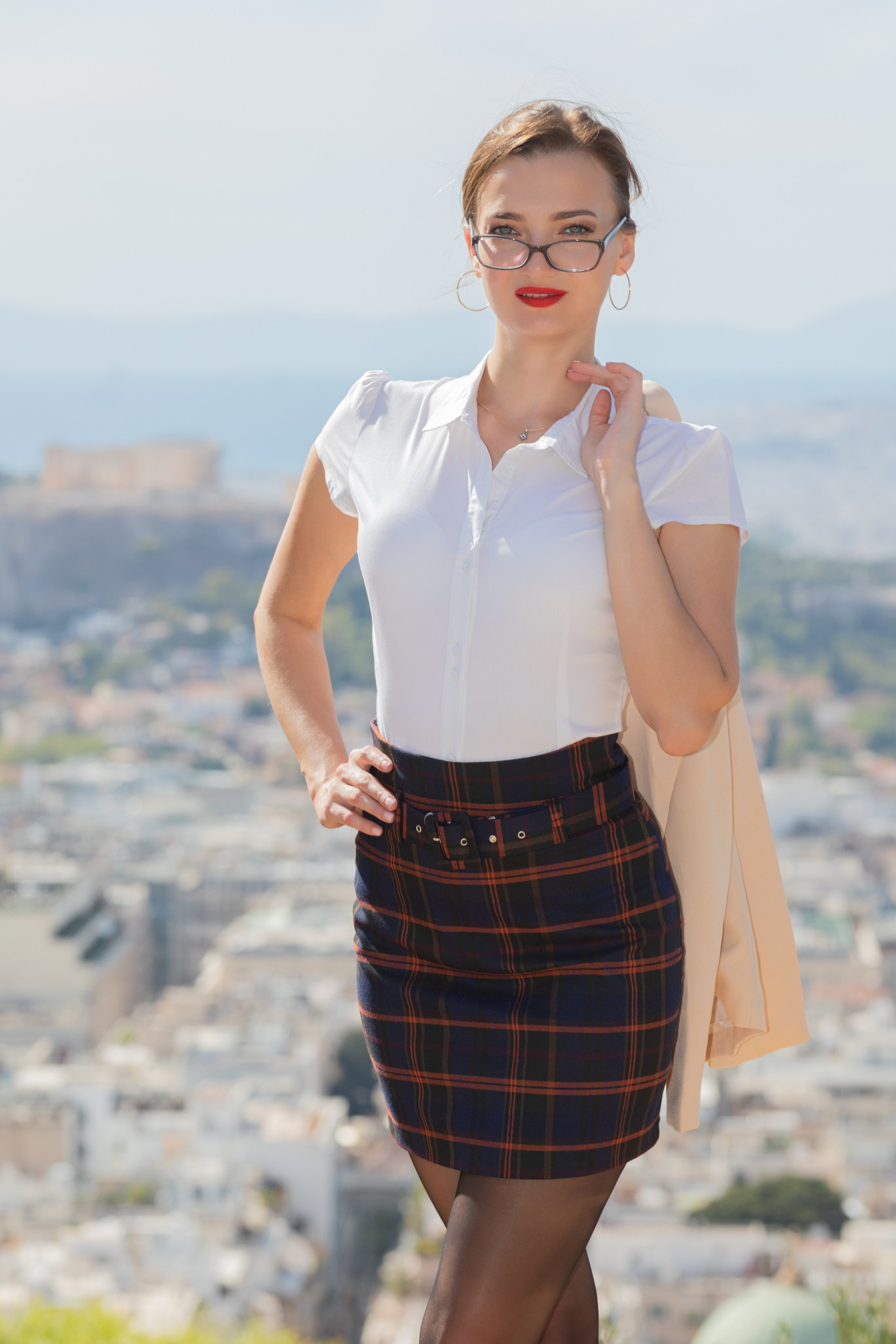 A woman with glasses and red lipstick wearing a white blouse and a dark plaid mini skirt, posing on a hill overlooking the city of Athens, with the Acropolis and Parthenon visible in the background.