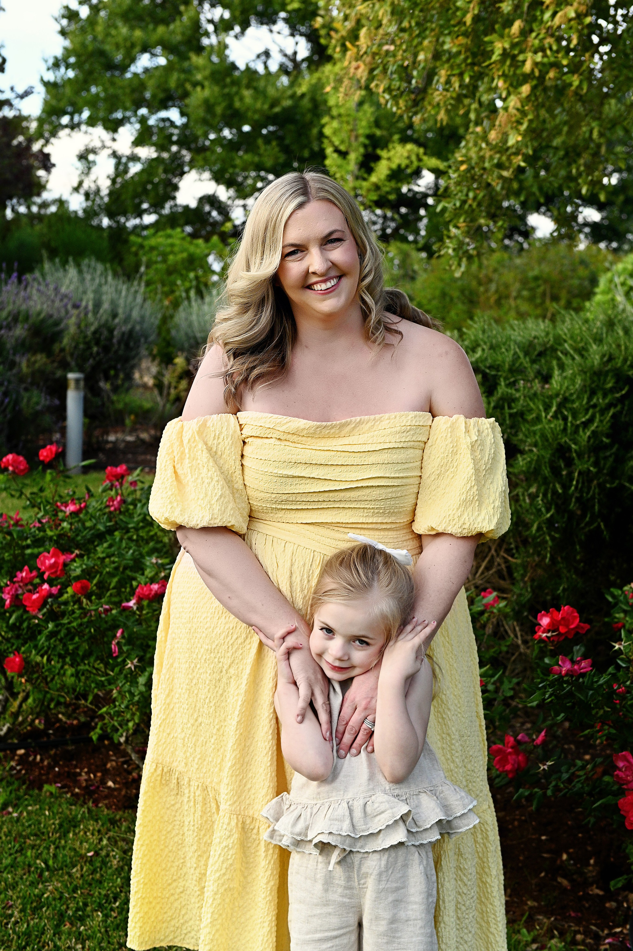 Fotografia di famiglia in Puglia: una donna sorridente in un abito giallo con spalle scoperte abbraccia una bambina in un completo crema, sullo sfondo di cespugli di rose rosse.