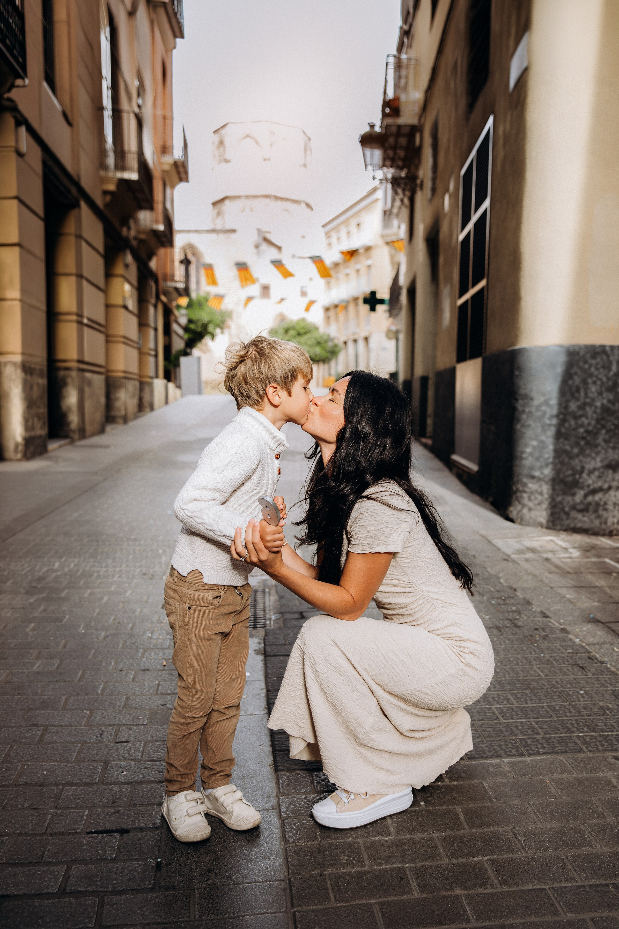 Sesión de fotos familiar en Valencia, España, con una madre cariñosa arrodillándose para besar a su pequeño hijo en una encantadora calle del casco histórico decorada con tradicionales banderas españolas. Un emotivo retrato lifestyle de madre e hijo que transmite cariño, recuerdos de la infancia y una auténtica conexión familiar. Ideal para fotografía familiar, sesiones de fotos familiares, retratos de madre e hijo, fotografía infantil, fotografía de vacaciones y fotografía familiar de destino en Valencia y toda España.