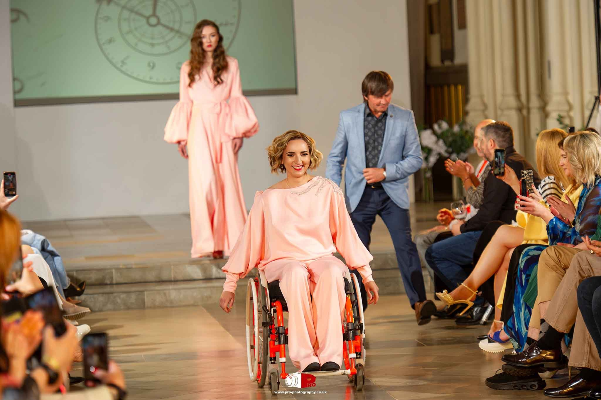 A smiling model in a wheelchair wearing a pink gown walks the runway at an inclusive fashion event while the audience applauds.
