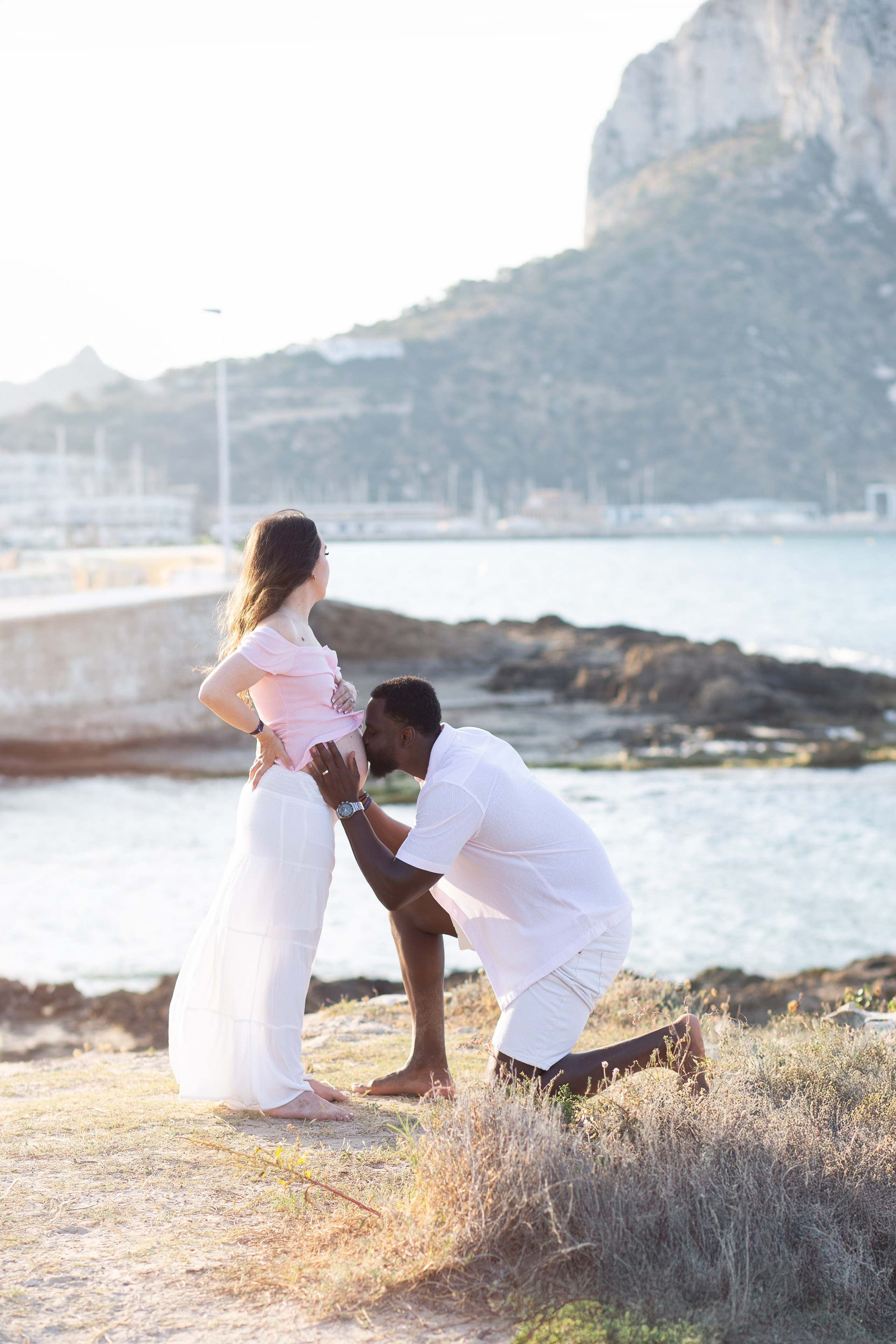 Sesión romántica de maternidad en Calpe, España — padre besando la barriga de su pareja con el Peñón de Ifach al fondo, capturando amor, ternura y expectación. Inspiración perfecta para sesiones escénicas de embarazo en la costa de Calpe.