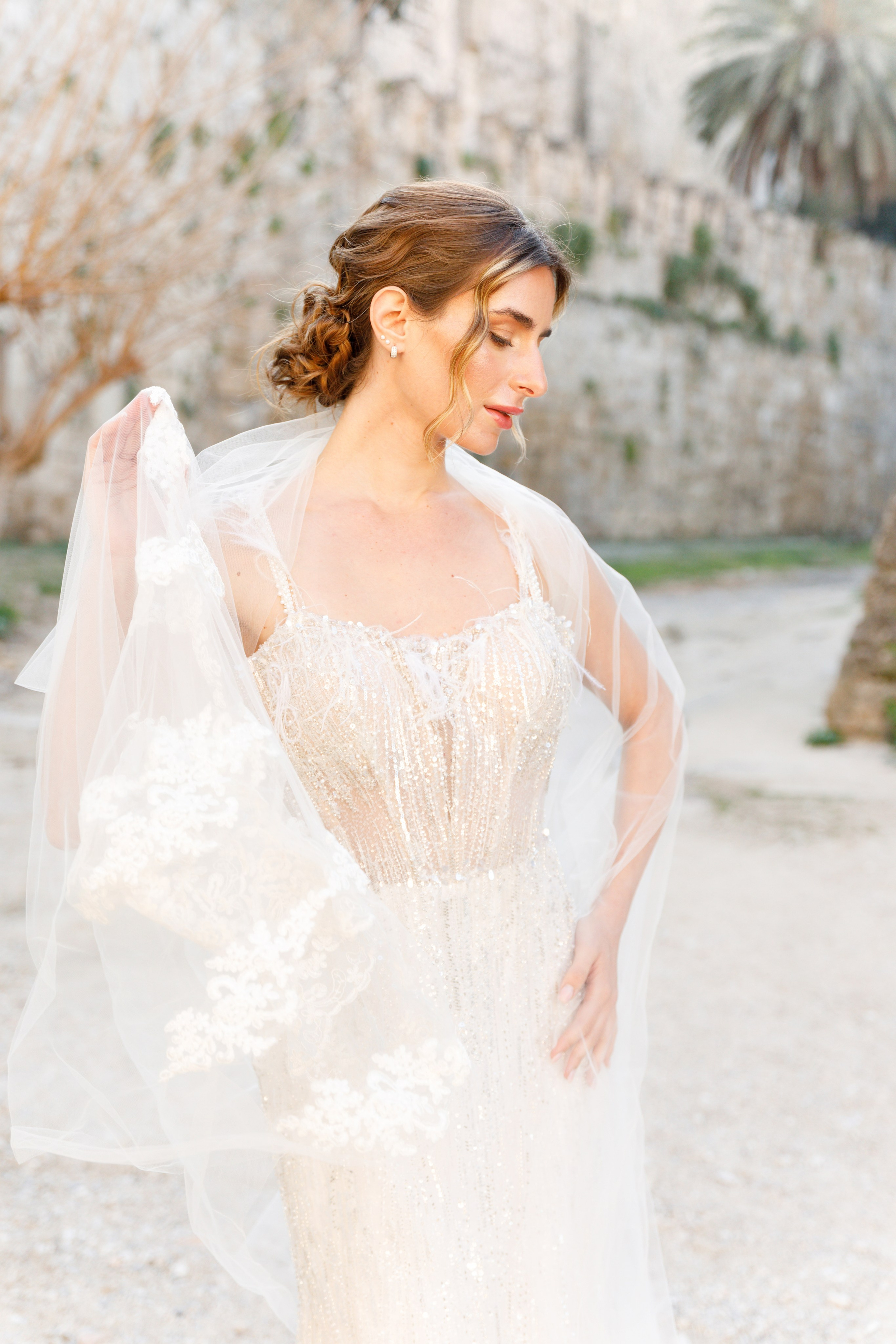 A radiant bride stands in the narrow, sunlit streets of Rhodes' Old Town, her intricate lace wedding dress contrasting beautifully with the weathered stone walls behind her. The editorial-style portrait highlights her serene expression and the timeless elegance of the historic setting, with soft natural light accentuating her features.