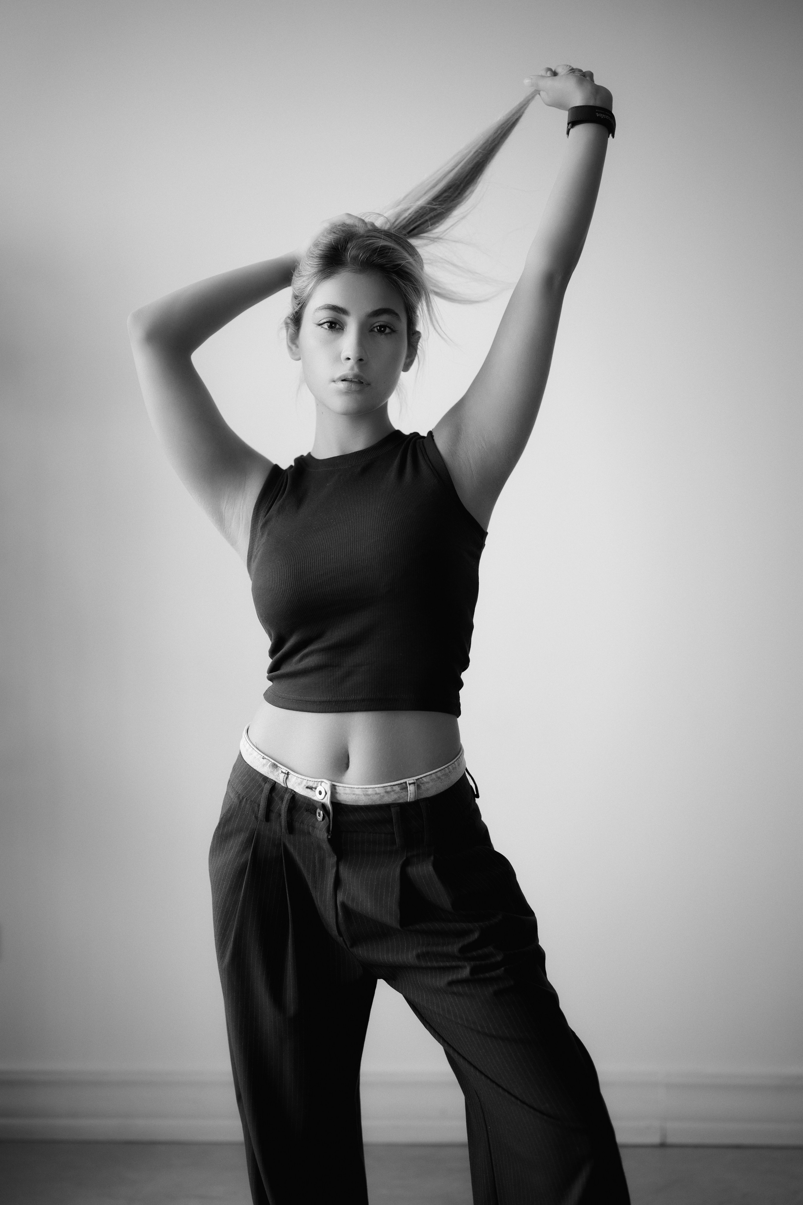 Black and white editorial fashion portrait of a woman standing with raised arms and holding her hair, photographed by Mehran Hadad with minimalist styling and soft light.