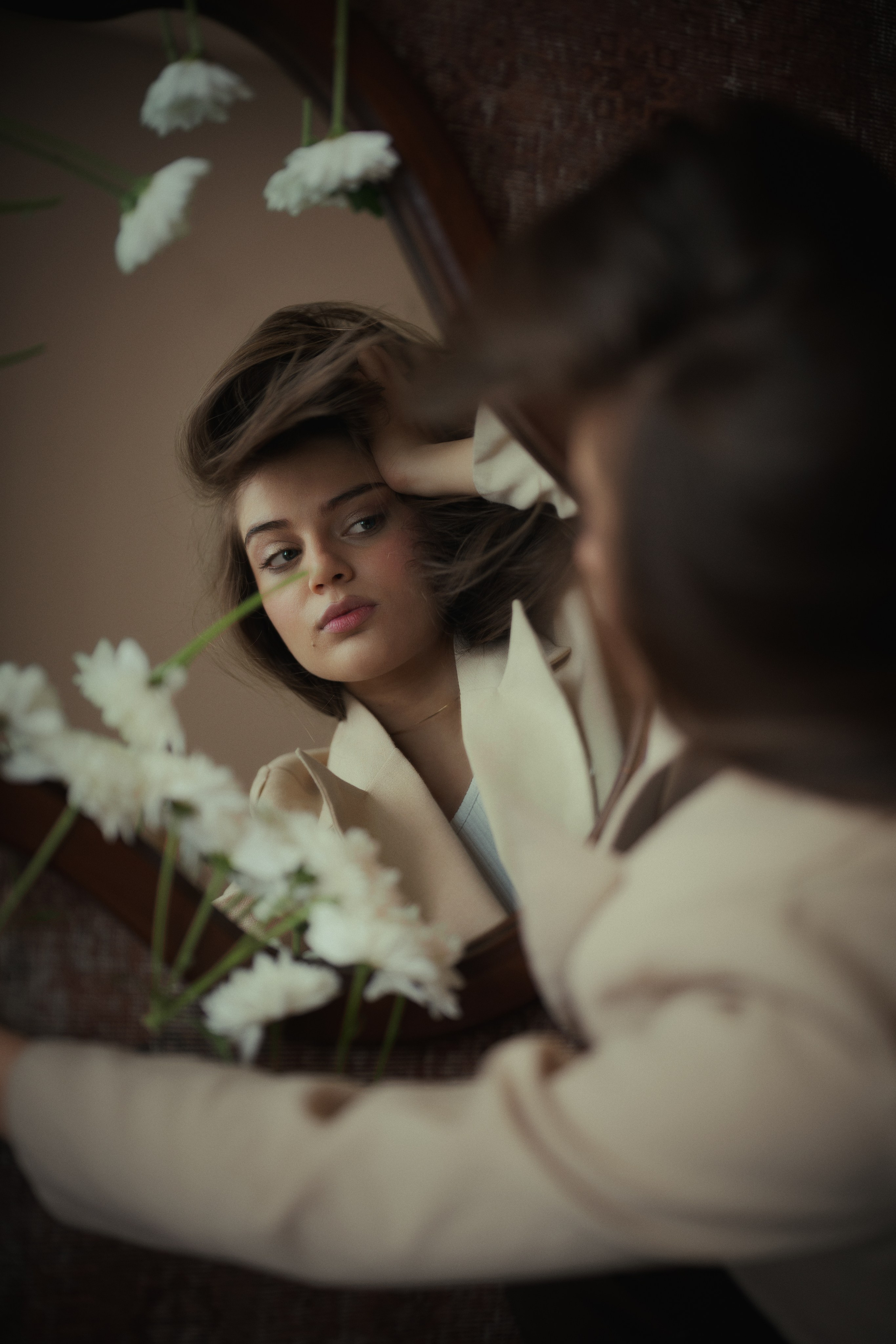 Cinematic mirror portrait of a woman in a beige suit surrounded by white flowers, photographed by Mehran Hadad with soft natural light.