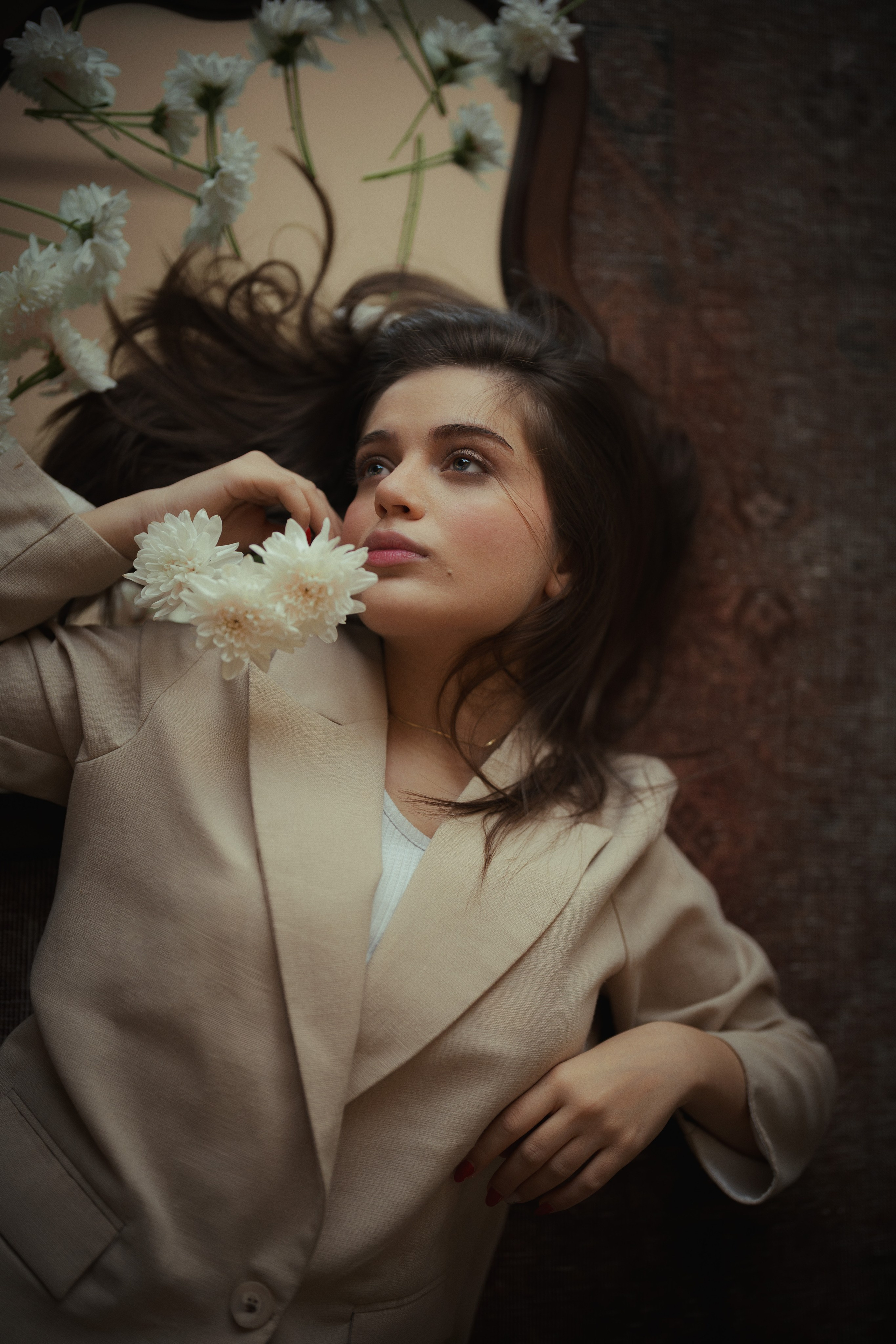Cinematic fashion portrait of a woman in a beige suit reclining beside a mirror and white flowers, photographed by Mehran Hadad.