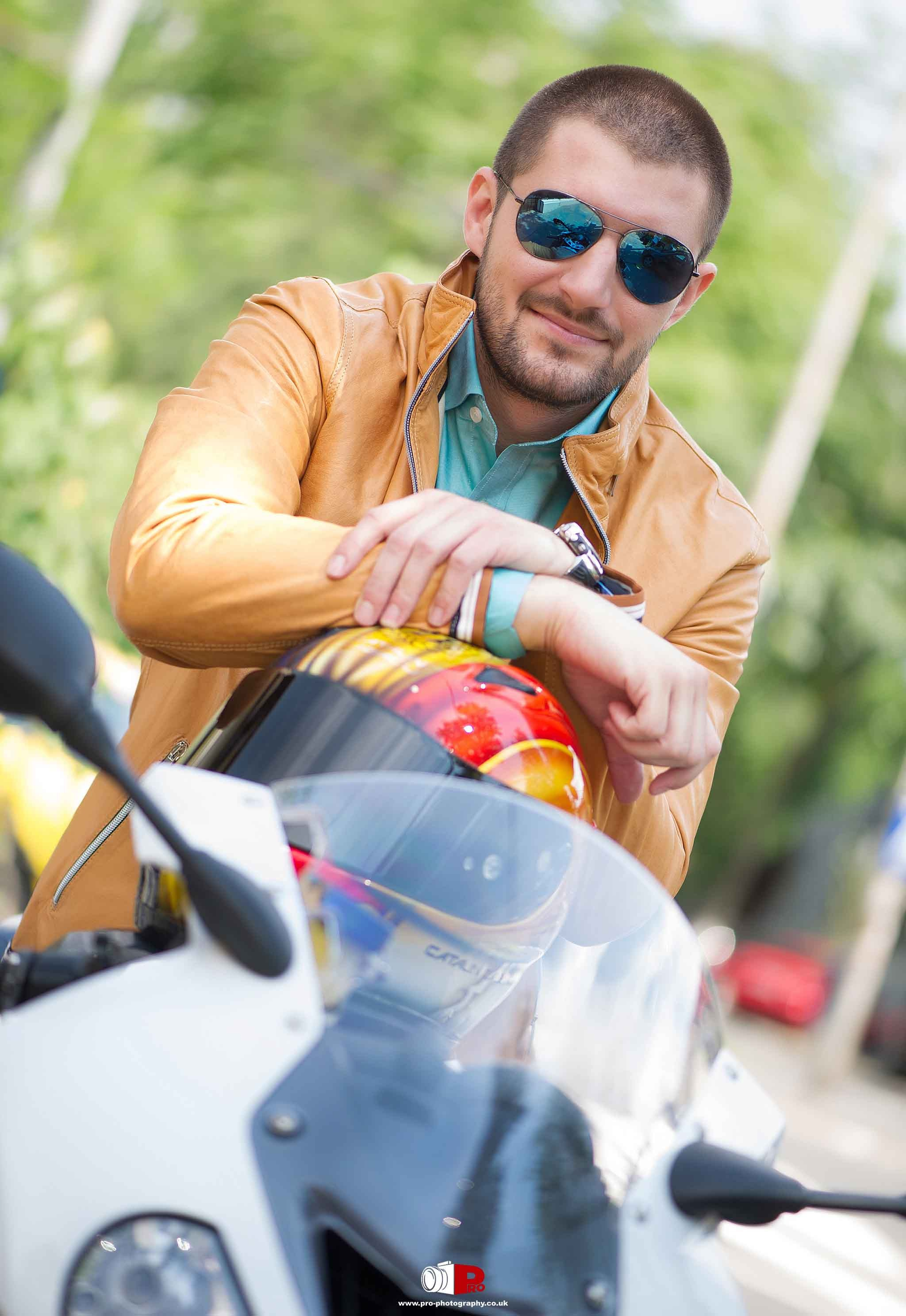 A man in a leather jacket and sunglasses posing beside his custom-designed motorcycle helmet.