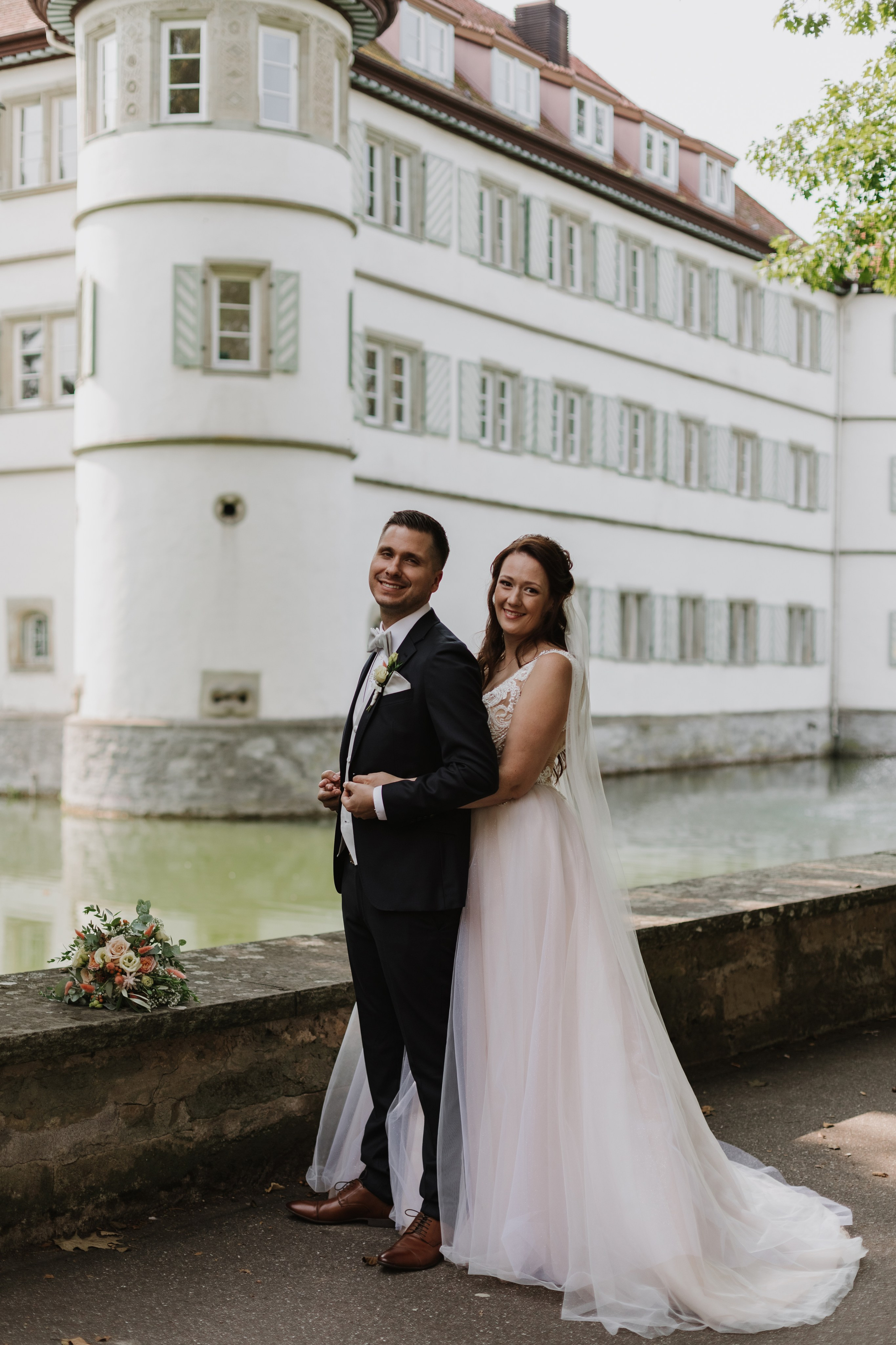 Braut umarmt Bräutigam von hinten vor einer Burgkulisse.