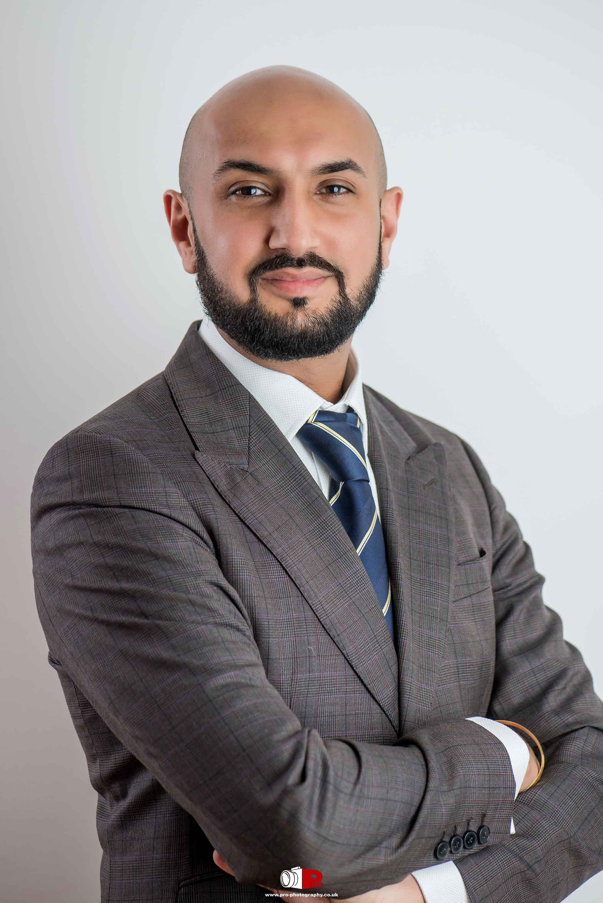 A confident bald man wearing a grey suit and striped tie posing for a corporate headshot against a white background.