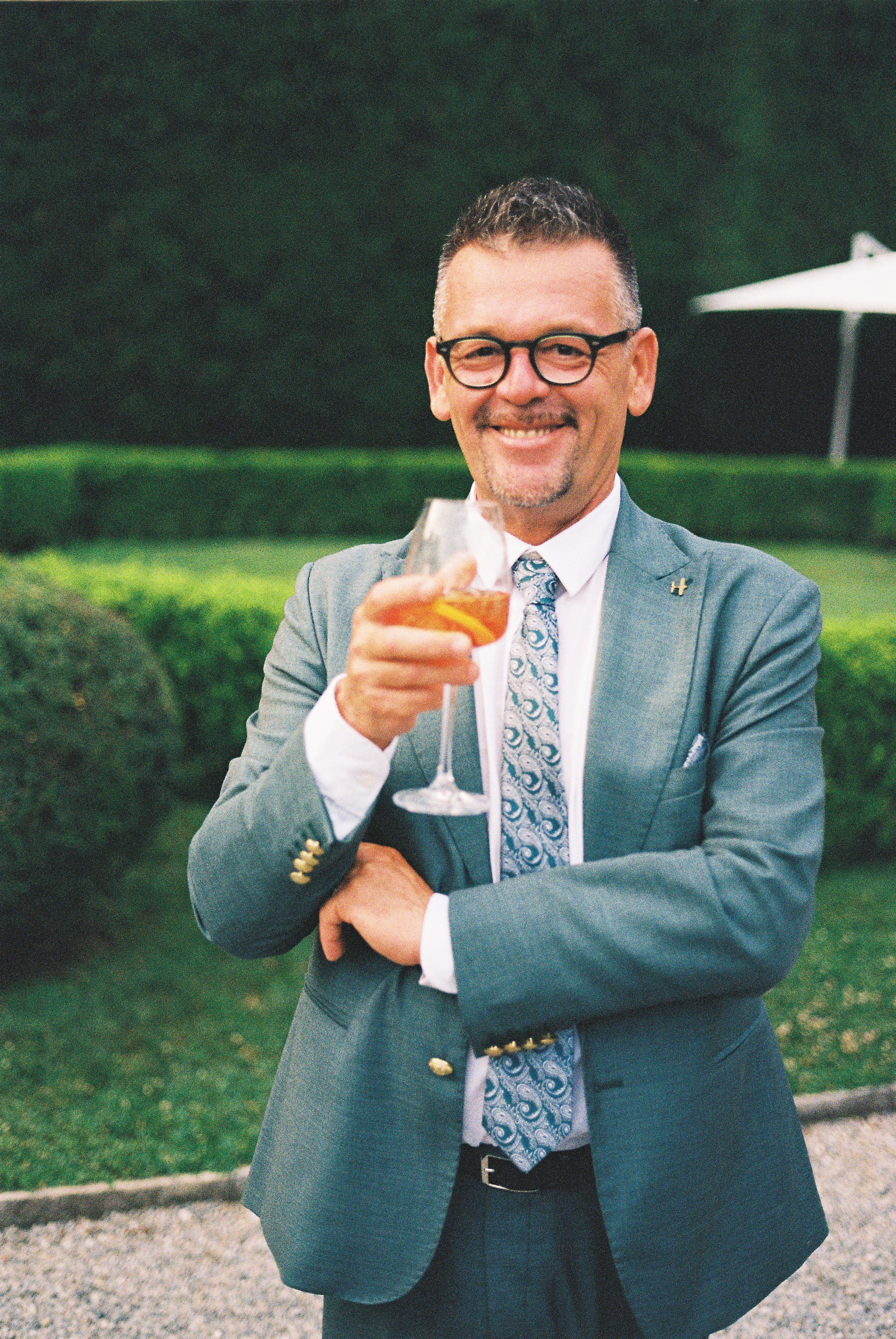 Man in a stylish grey suit smiling, holding a glass of orange drink outdoors.