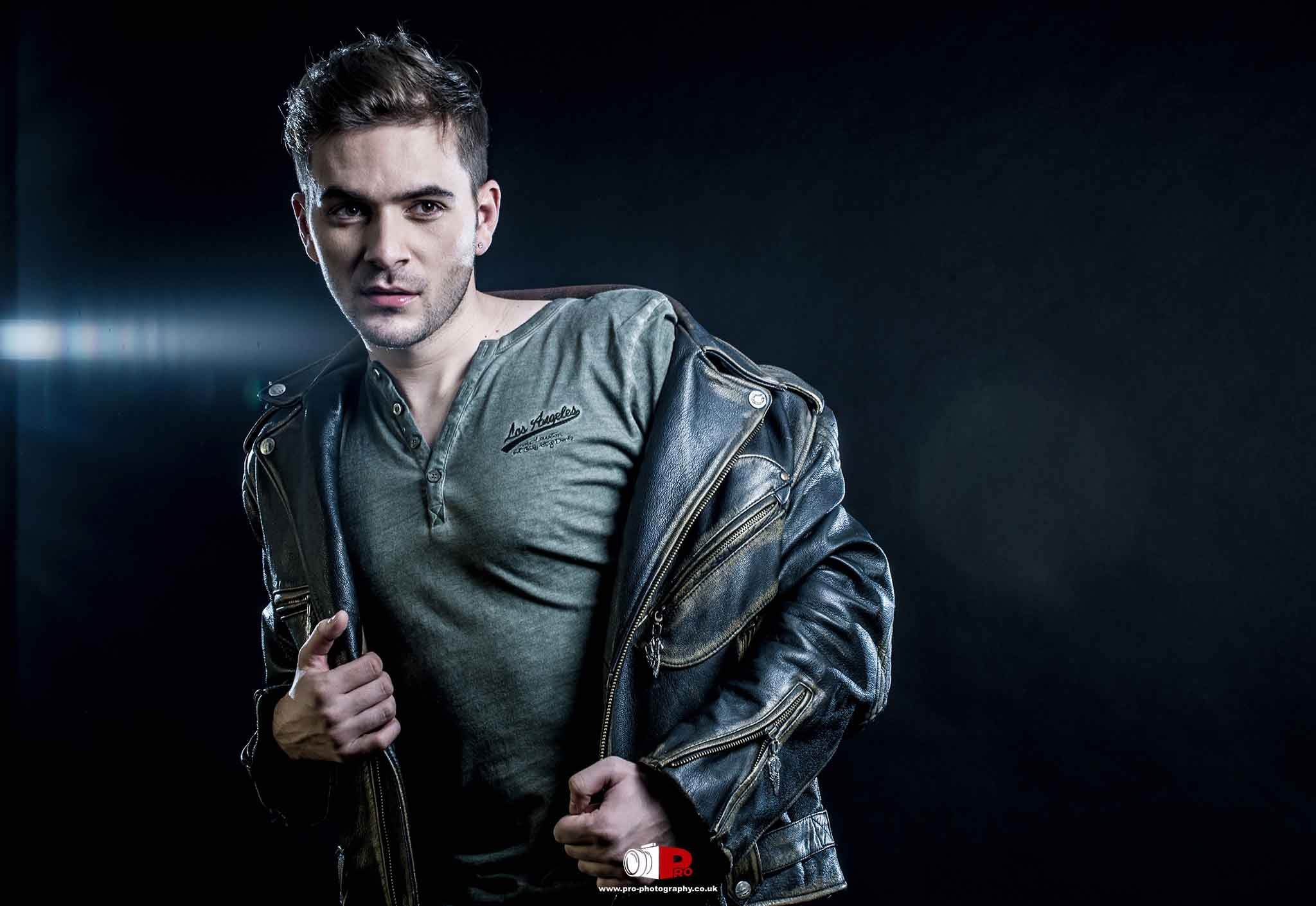 A man in a grey shirt and leather jacket posing in a moody, cinematic studio portrait.
