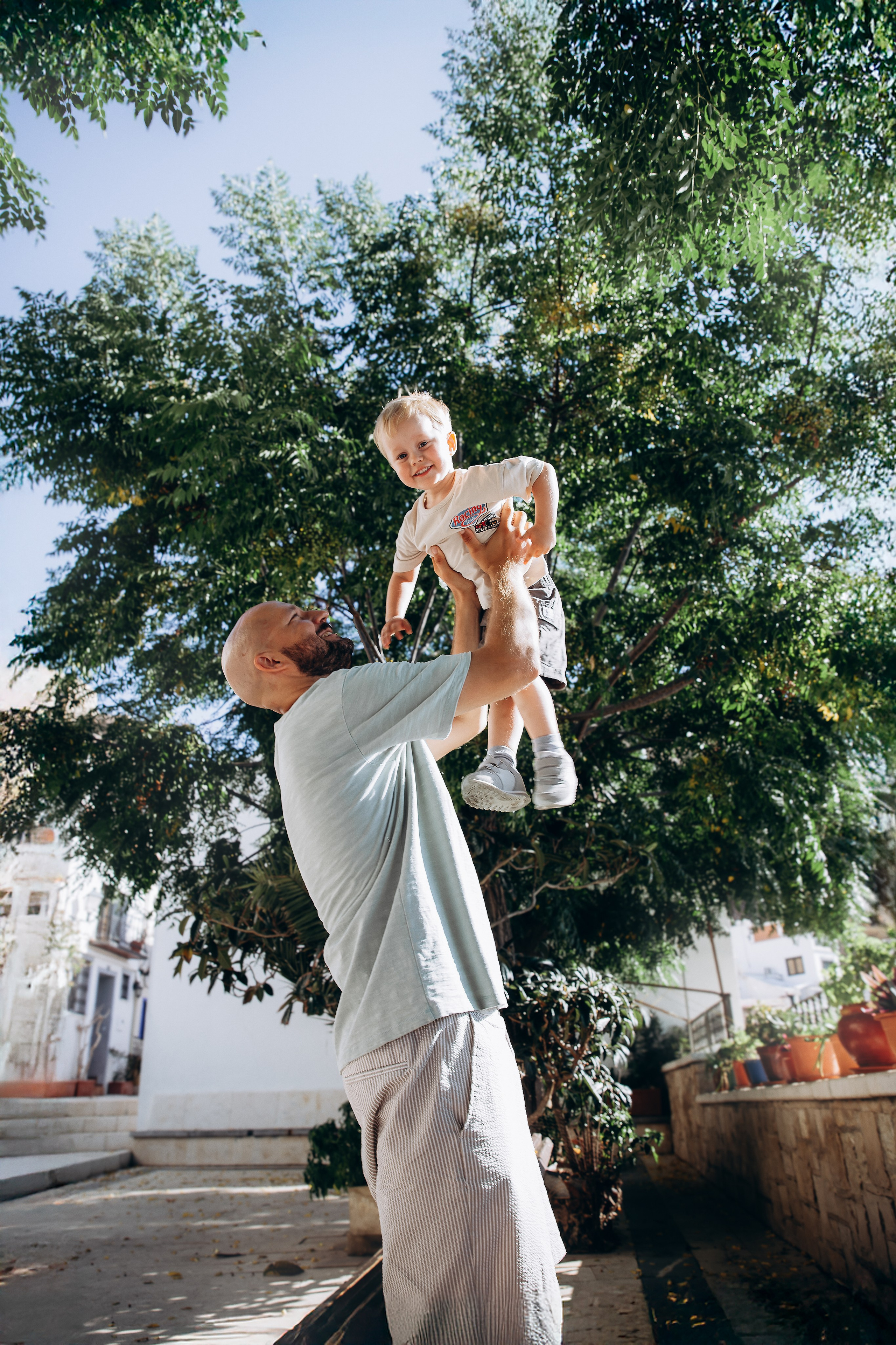 Foto familiar entrañable tomada en Valencia, España, con un padre levantando con alegría a su hijo pequeño bajo árboles frondosos — perfecta para familias que desean conservar momentos naturales durante una sesión familiar en Valencia y España.