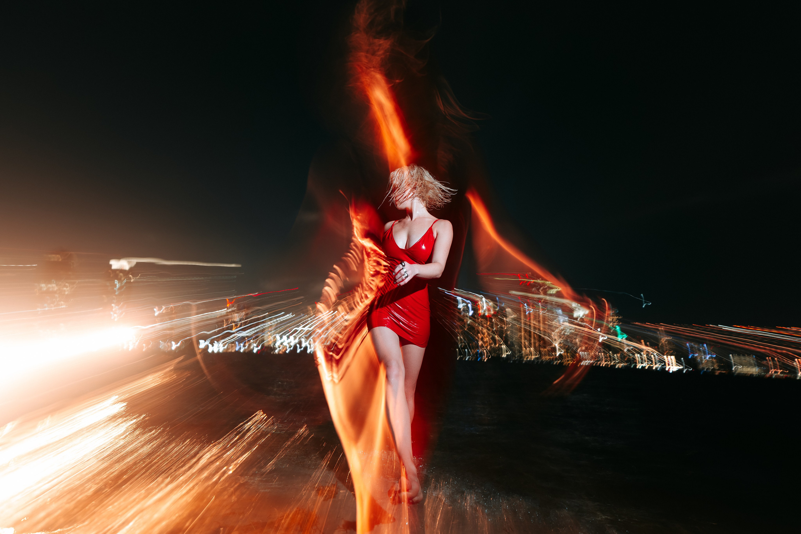 The red dress moves in the wind as the model stands barefoot by the water. Lighting highlights the fabric’s motion.