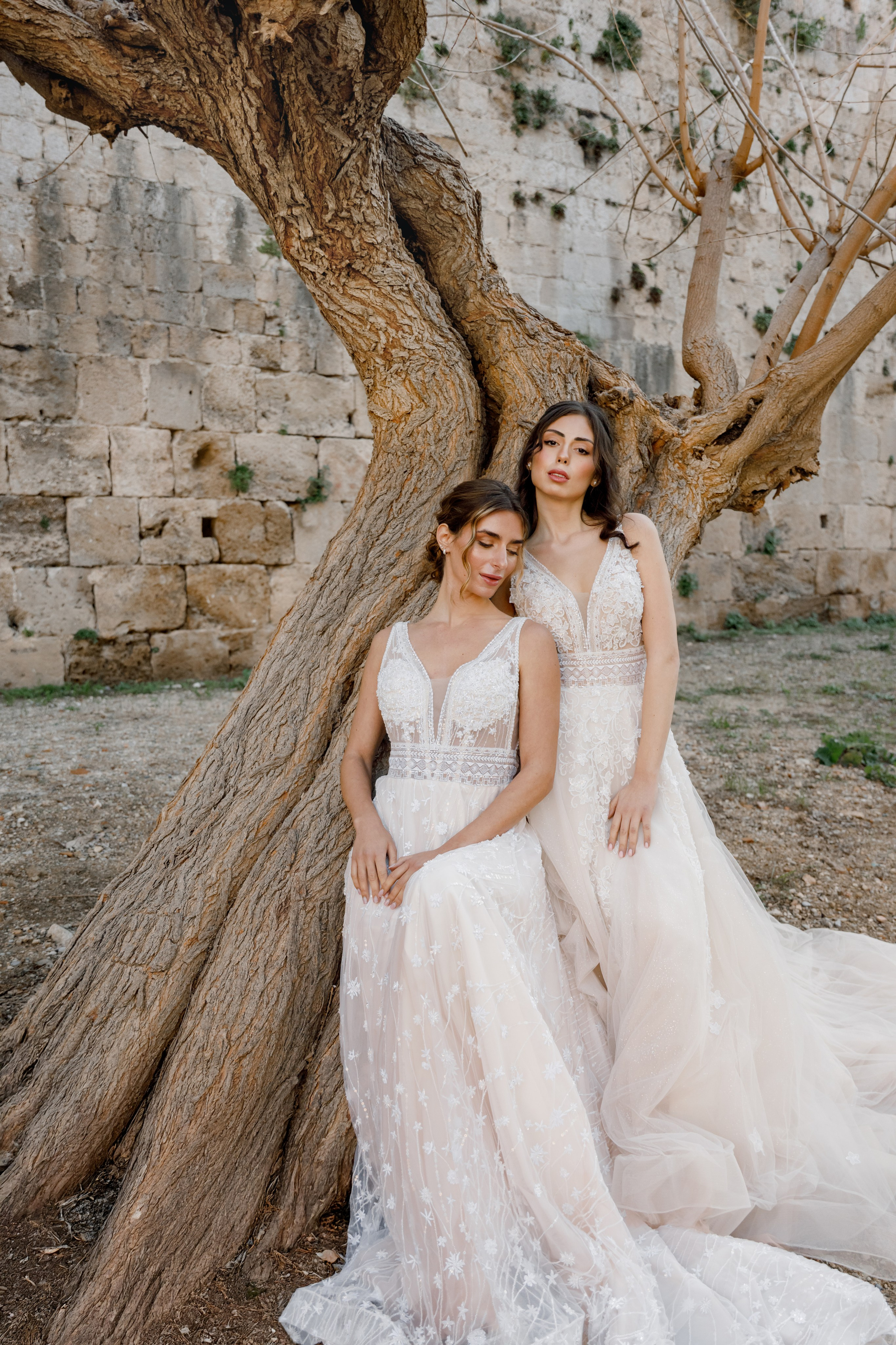 Two brides in elegant wedding dresses pose in the narrow, cobblestone streets of Rhodes' Old Town, surrounded by historic stone buildings and vibrant bougainvillea. The editorial-style photo captures their intimate connection and the timeless charm of the medieval setting, with soft natural light enhancing the romantic atmosphere.