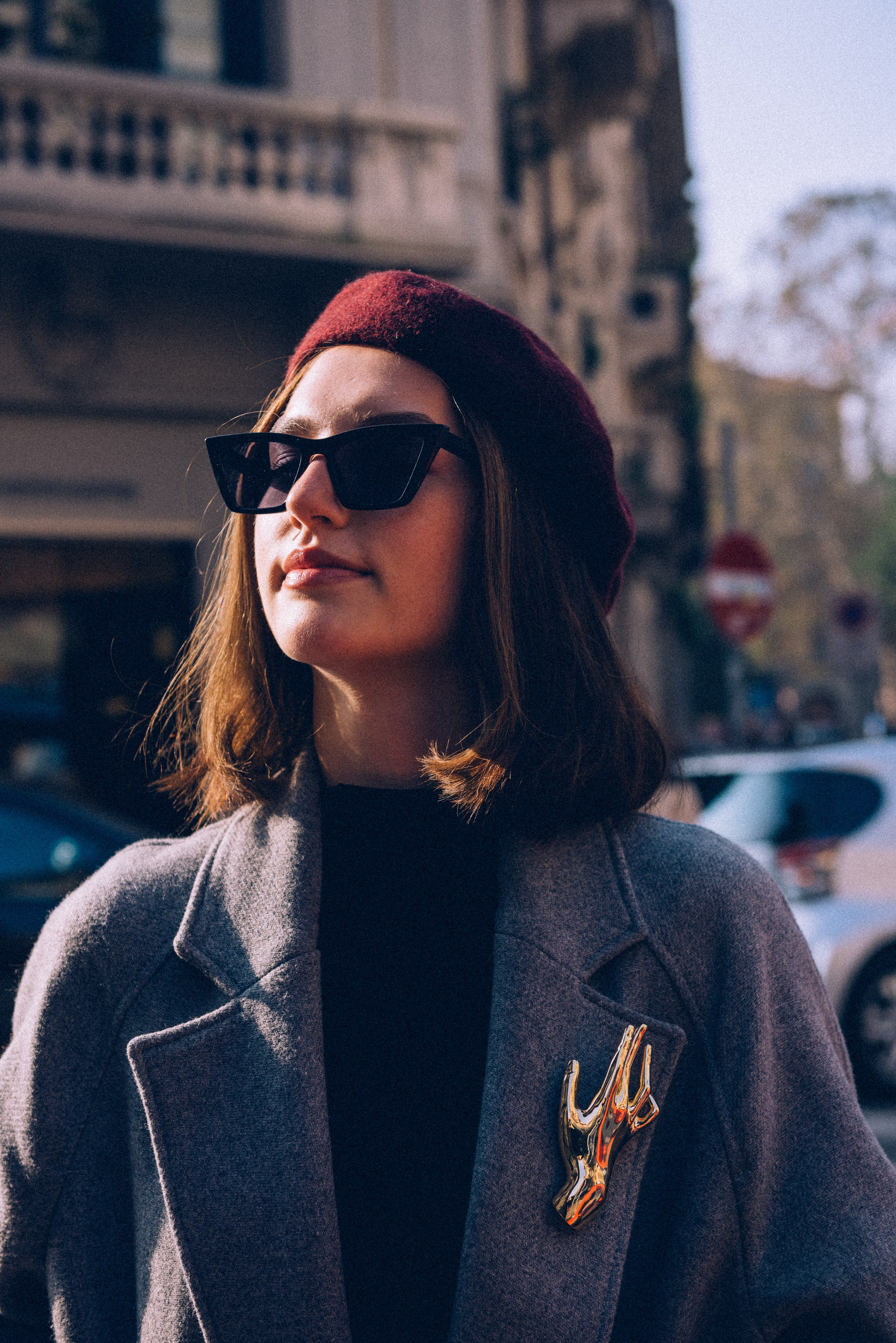 Close-up portrait of a stylish woman in Milan wearing sunglasses, a gray coat, and a vintage gold brooch under warm morning light