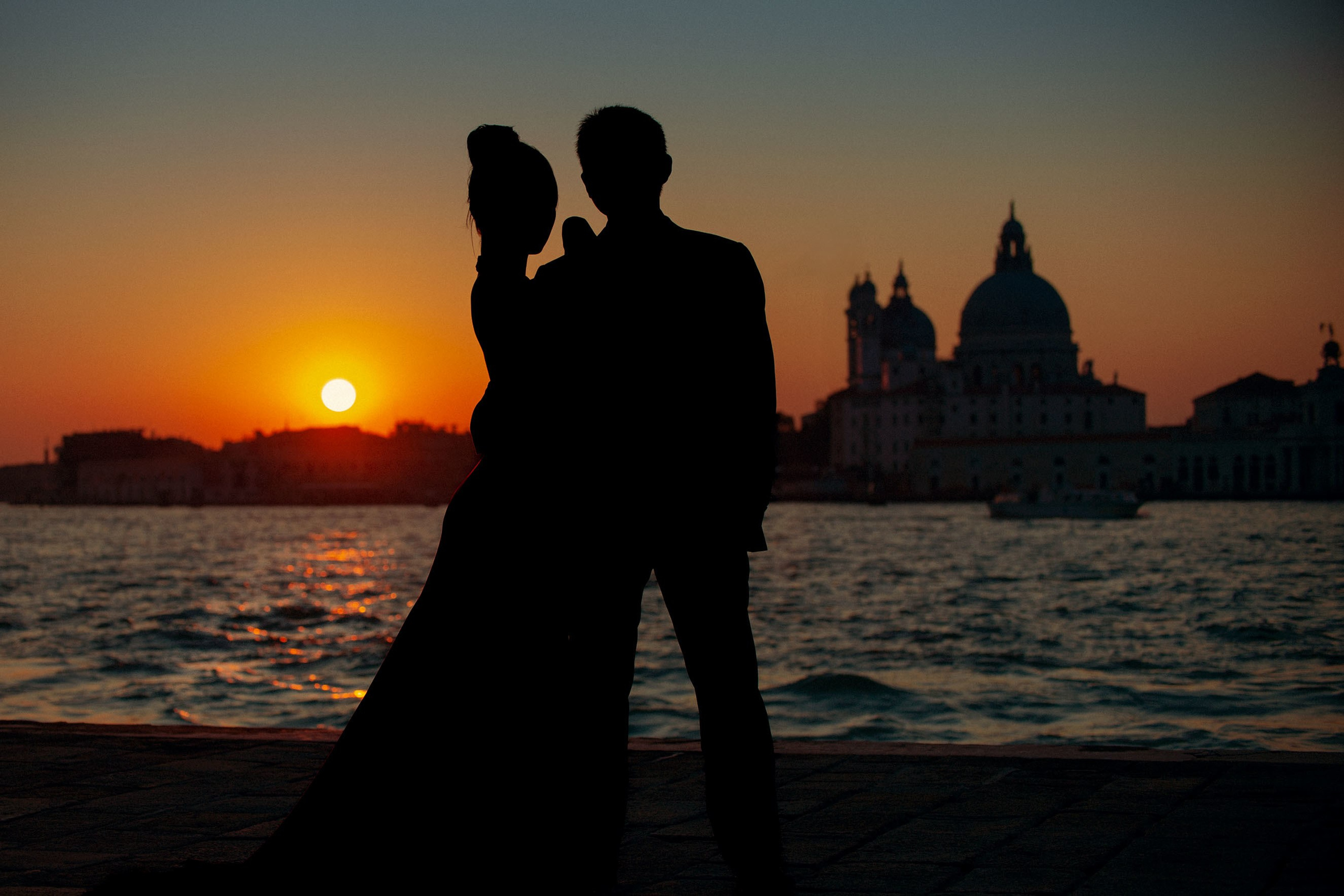 Couple in silhouette watching sunset over Venice's historic skyline near its canal.