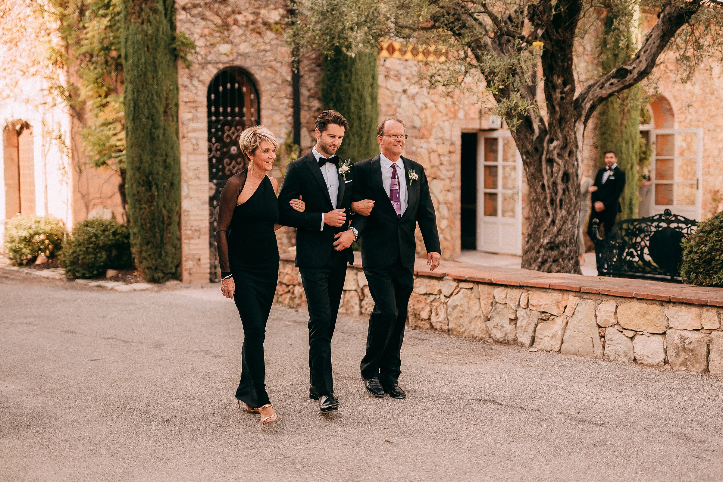 The groom walks arm in arm with his parents down a tree-lined path in Provence, France, with warm golden light illuminating their path.