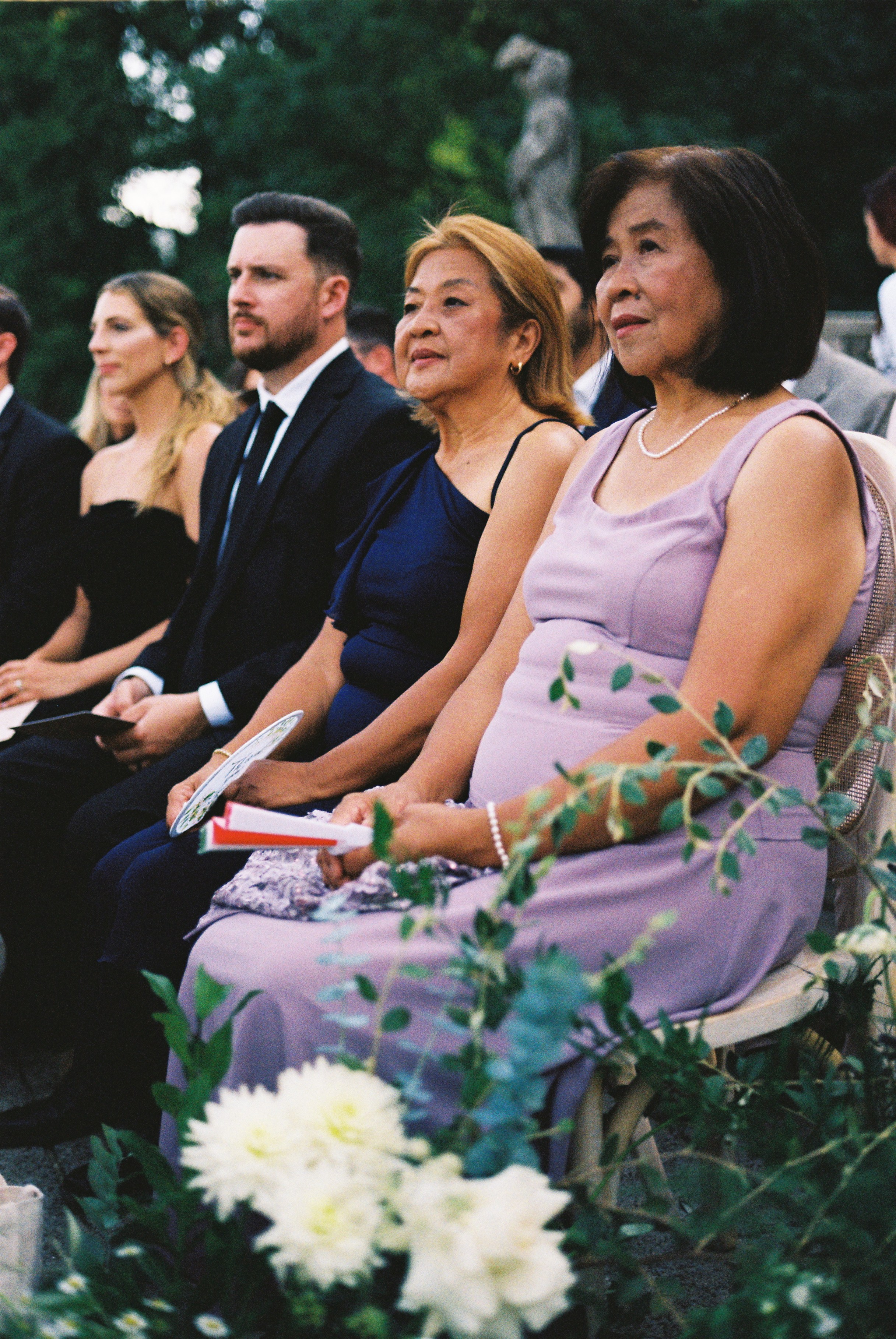 Family members in elegant attire sit in ceremony, surrounded by greenery and floral decorations.