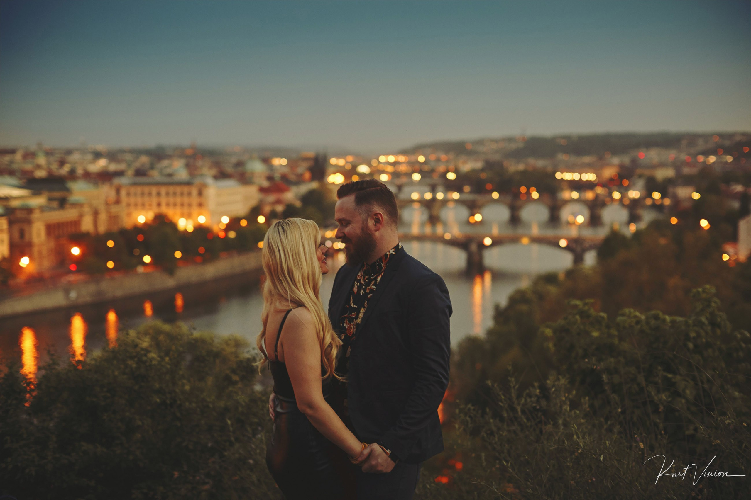 A blonde-haired woman embraces her stylish dark-haired partner as the twinkling lights of Prague at dusk are seen below them from their vantage point above the city