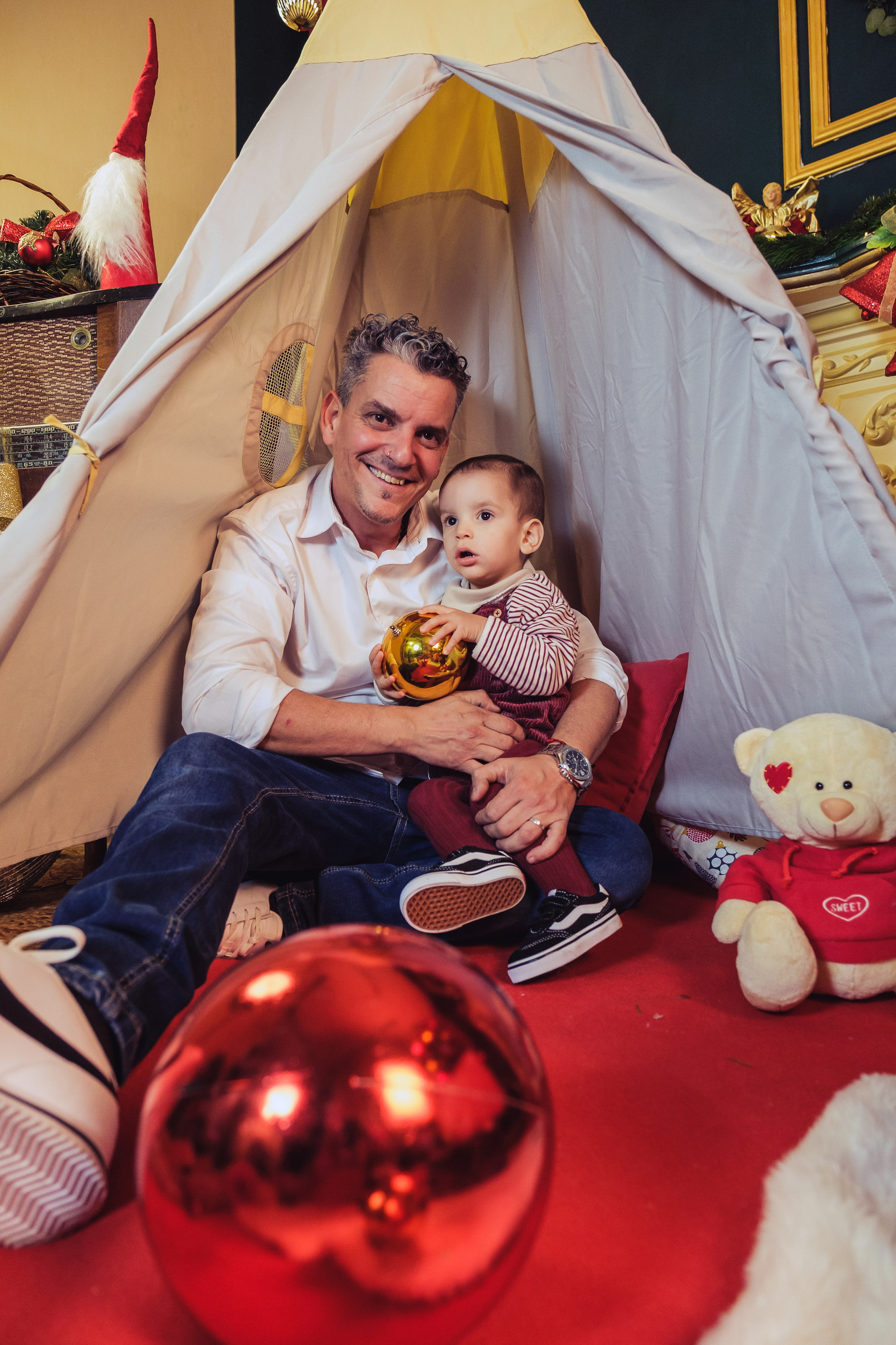 padre y bebé sentados dentro de una tienda de lona con bolas rojas y peluches, sesión de Navidad en estudio