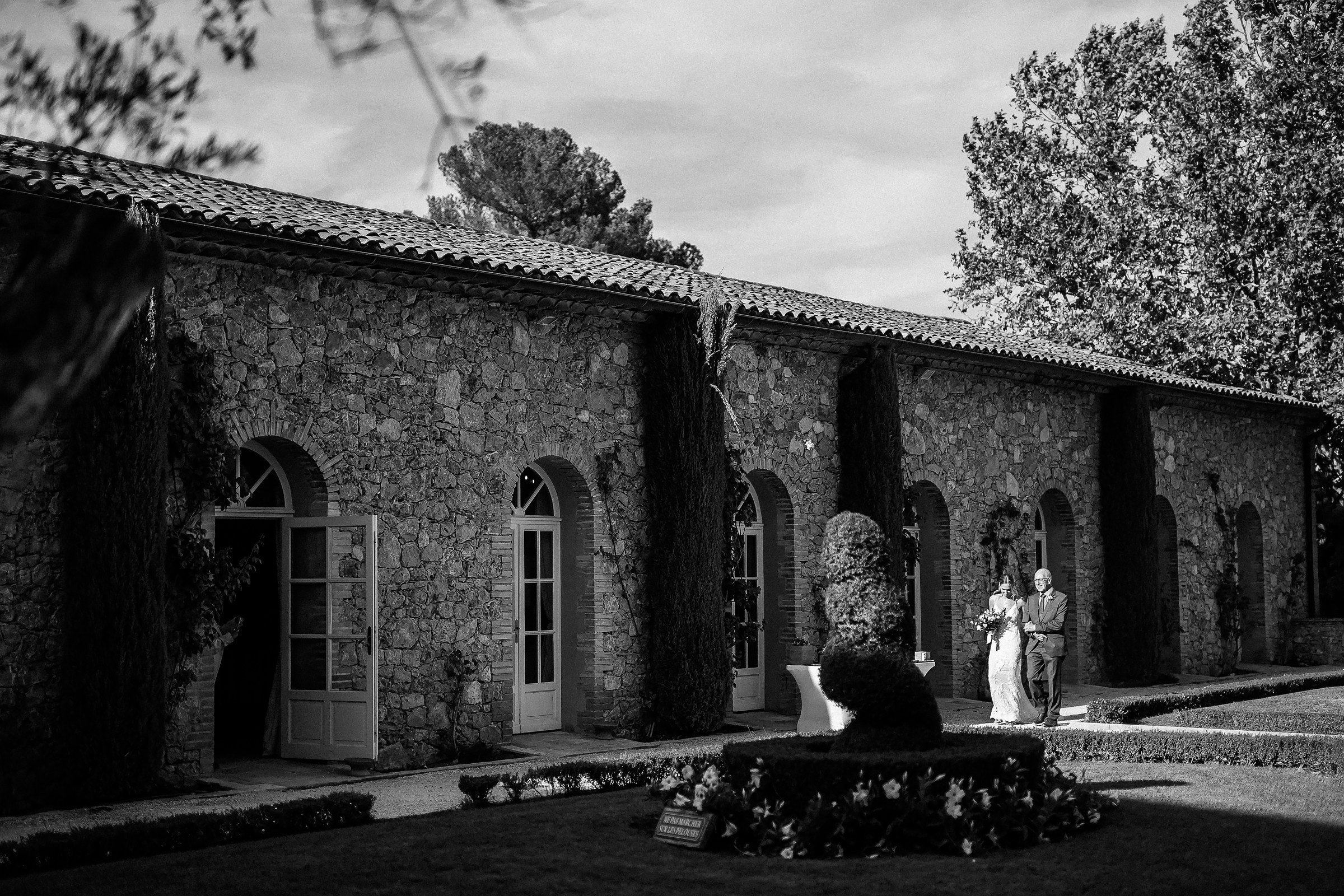 A wide black-and-white view of the bride with her parents standing in a manicured garden courtyard framed by tall trees and stone buildings in Provence, France.