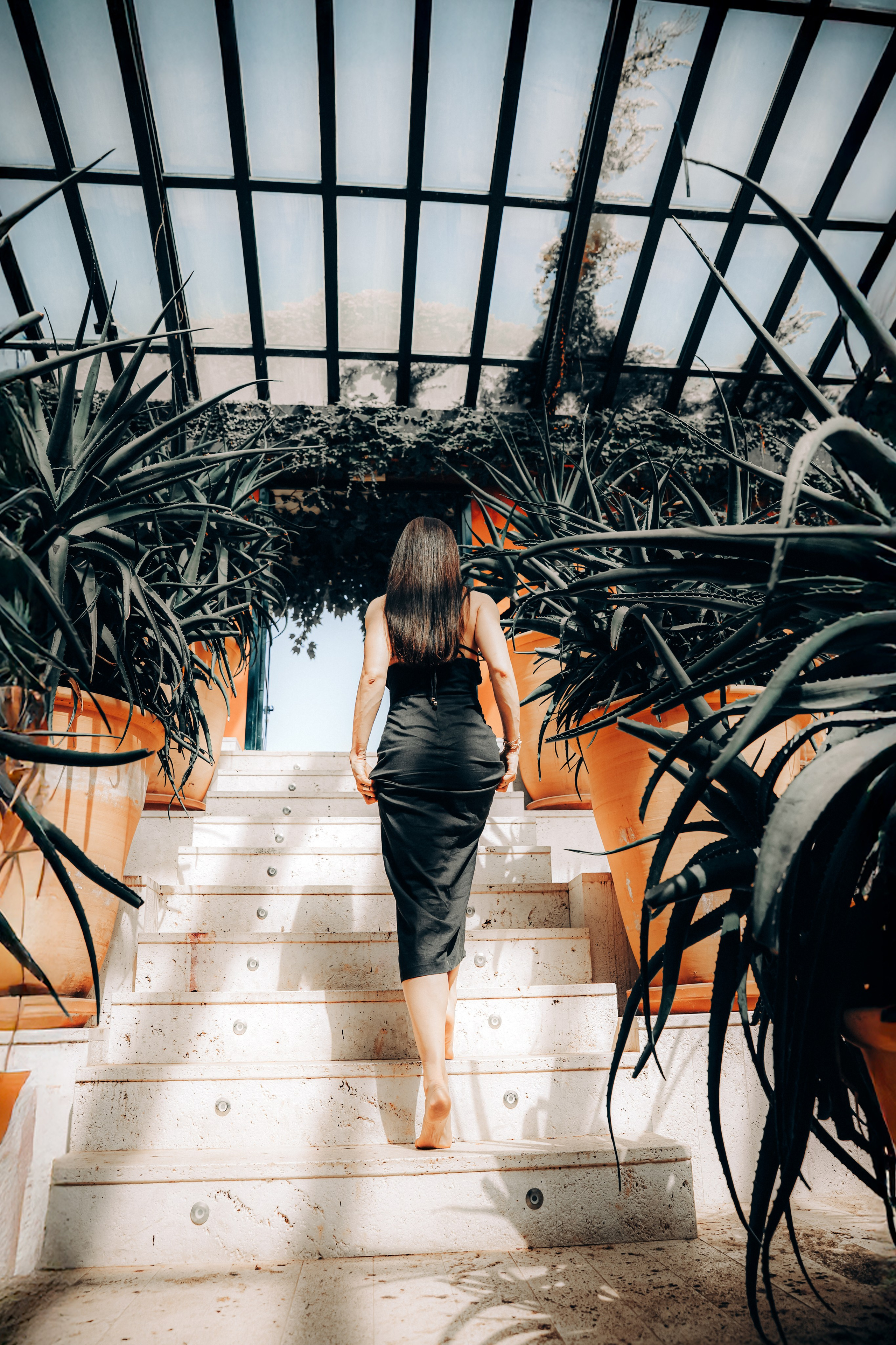 Женщина в элегантном черном платье идет босиком вверх по мраморной лестнице среди драматичных тропических растений в современном помещении с стеклянной крышей в Кальпе, Испания — стильная портретная фотосессия, сочетающая архитектурные линии и природные элементы, идеально подходит для портретной съемки в Кальпе и Испании.