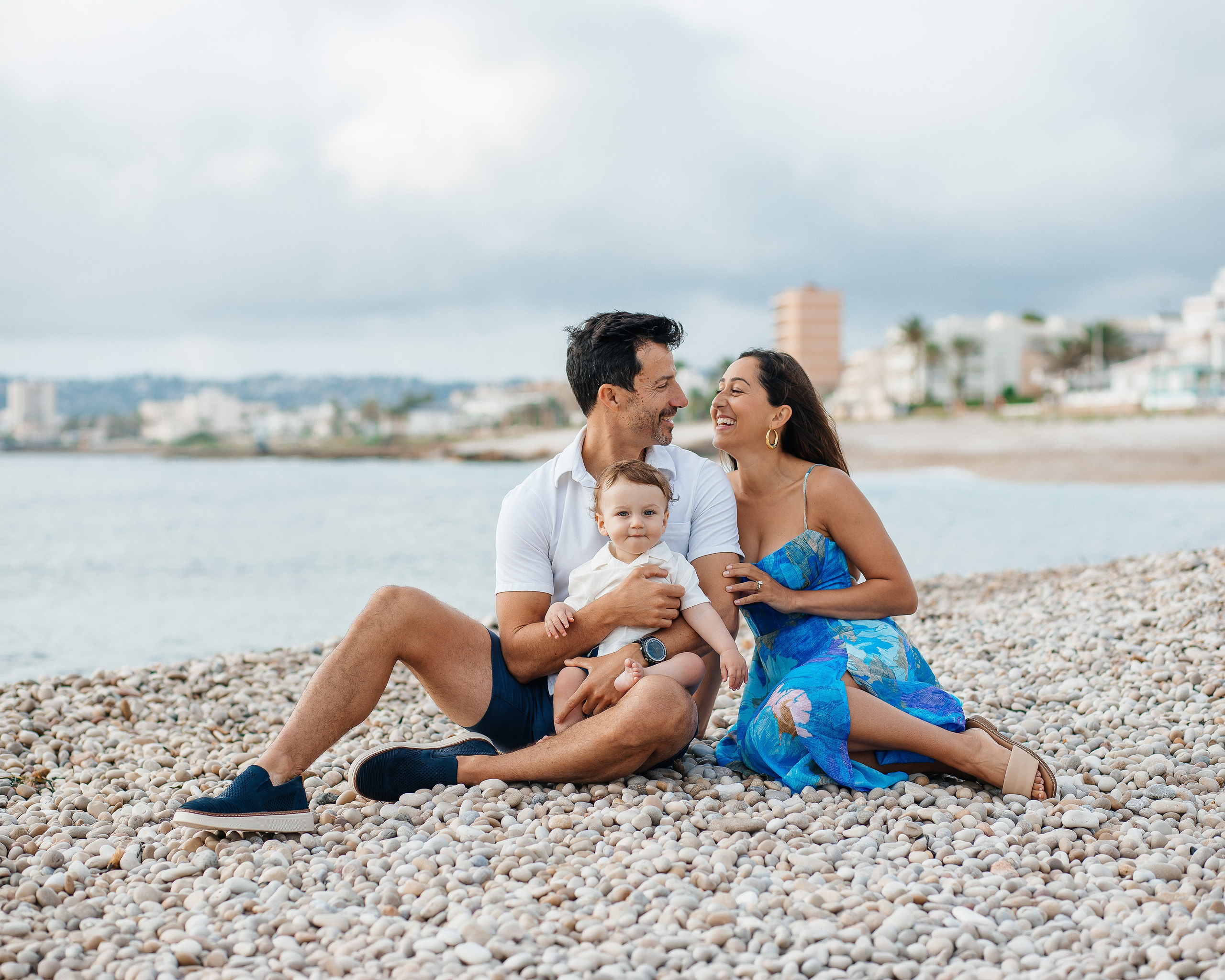 Sesión de fotos familiar en Valencia, España, con una feliz familia y su pequeño hijo sentados juntos en una playa del Mediterráneo, compartiendo sonrisas y momentos inolvidables junto al mar. Un retrato familiar lifestyle con vistas a la costa, ideal para fotografía familiar, sesiones familiares en la playa, fotografía de vacaciones y fotografía familiar de destino en Valencia y toda España.