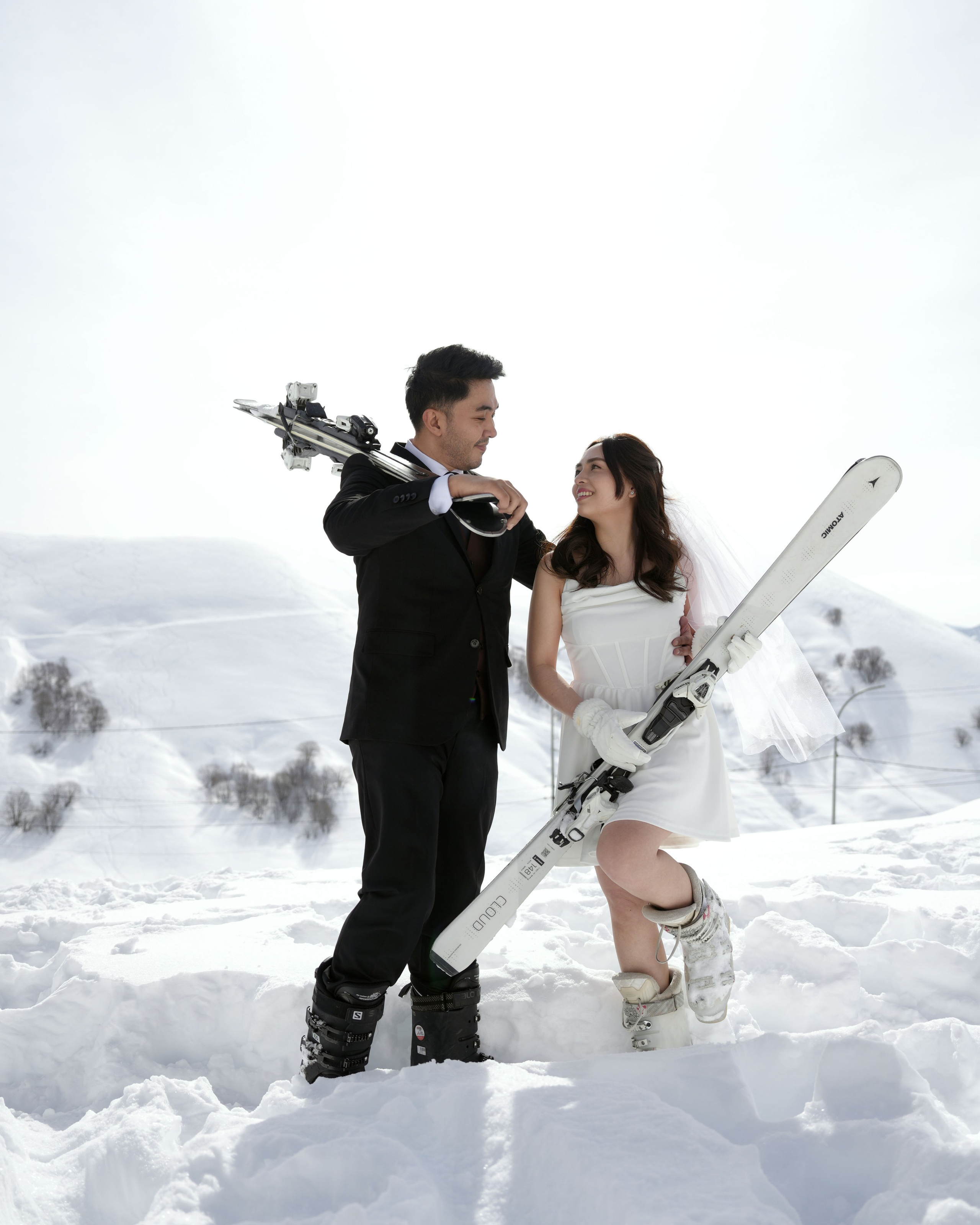 Bride in white jacket and groom in ski gear posing with skis in Gudauri Ski Resort
