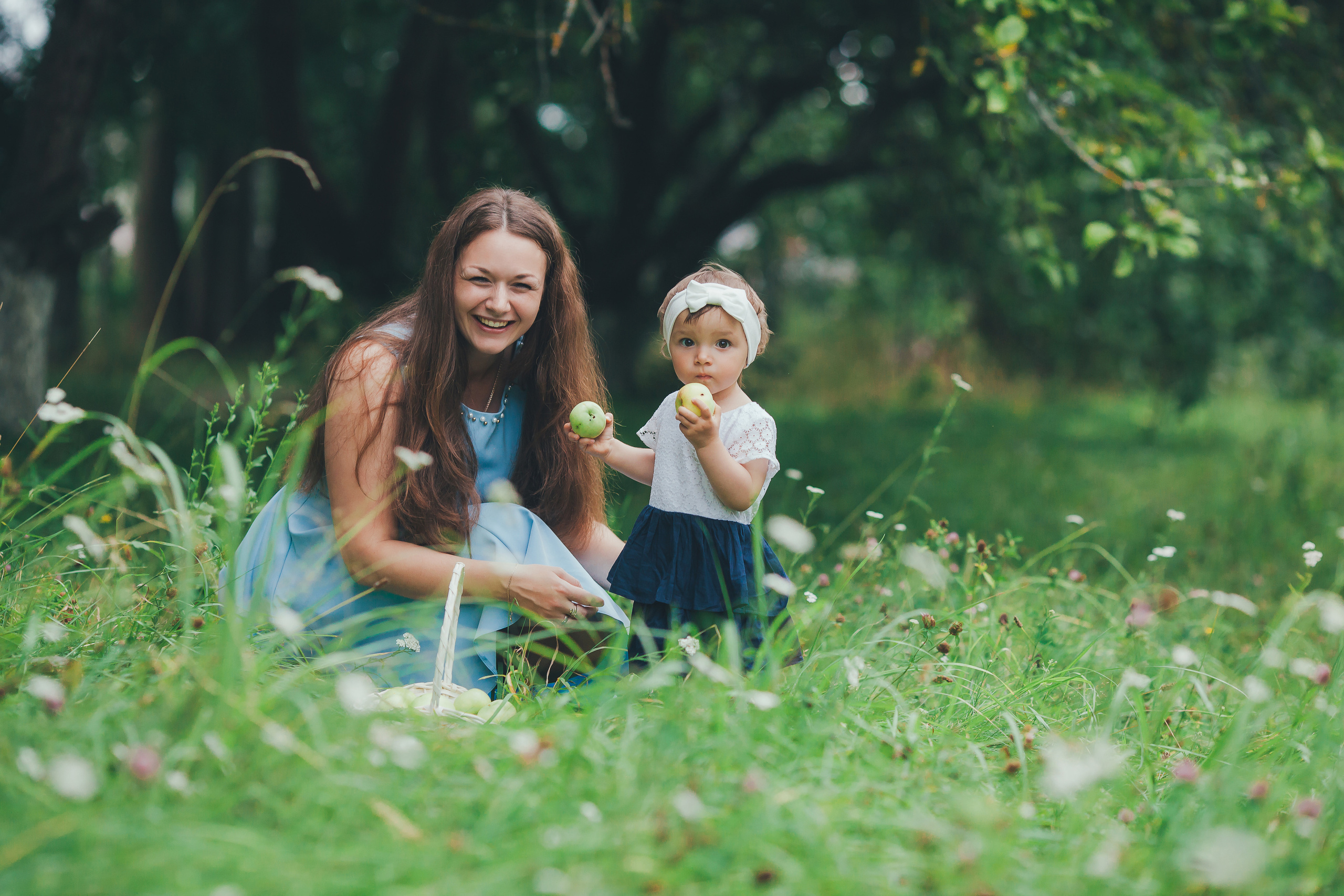Мама с дочкой сидят на полянке и кушают зеленые яблоки.