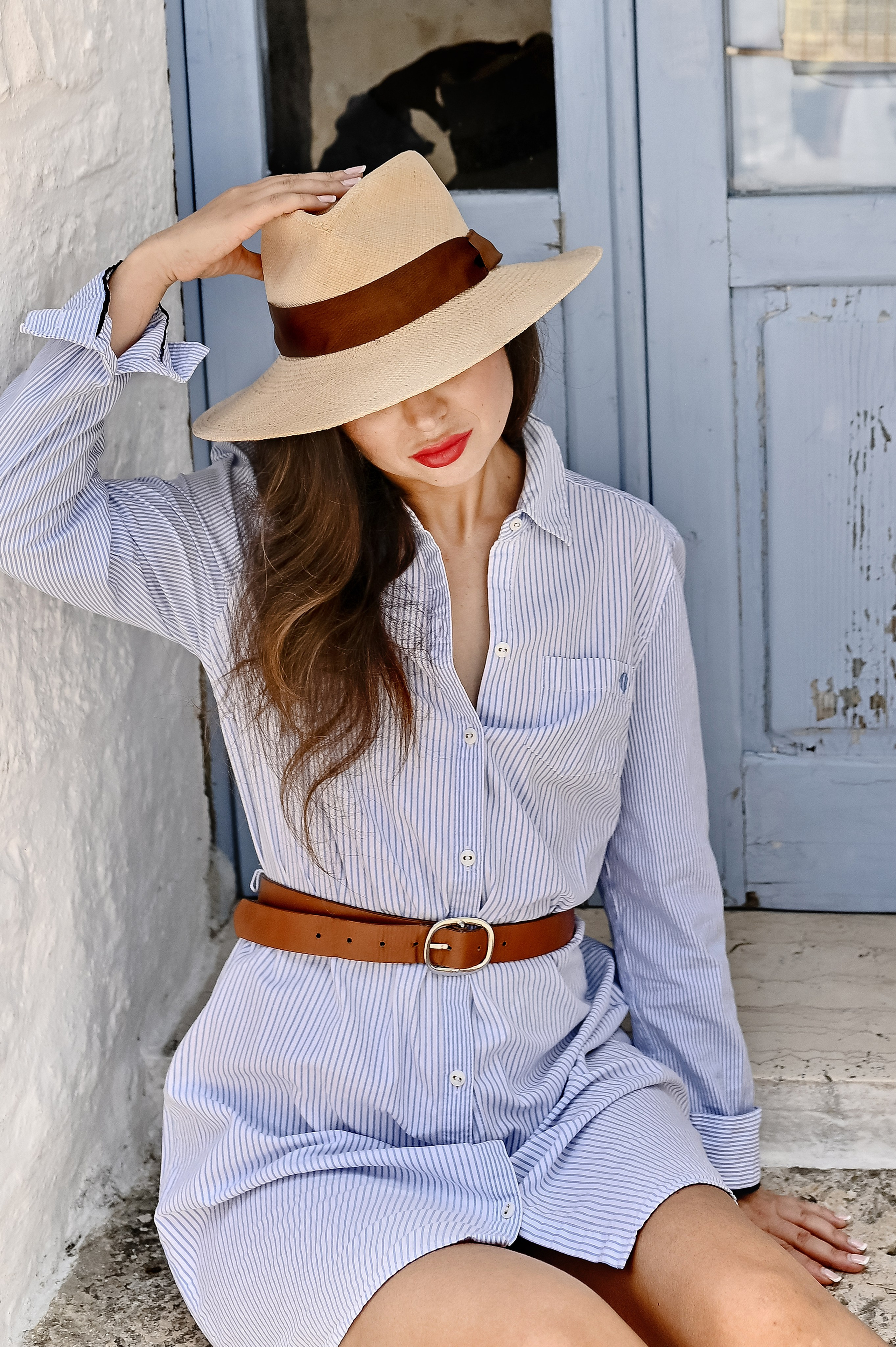 A woman in a light blue shirt dress and straw hat sits by a blue wooden door, tilting her head and shading her face.