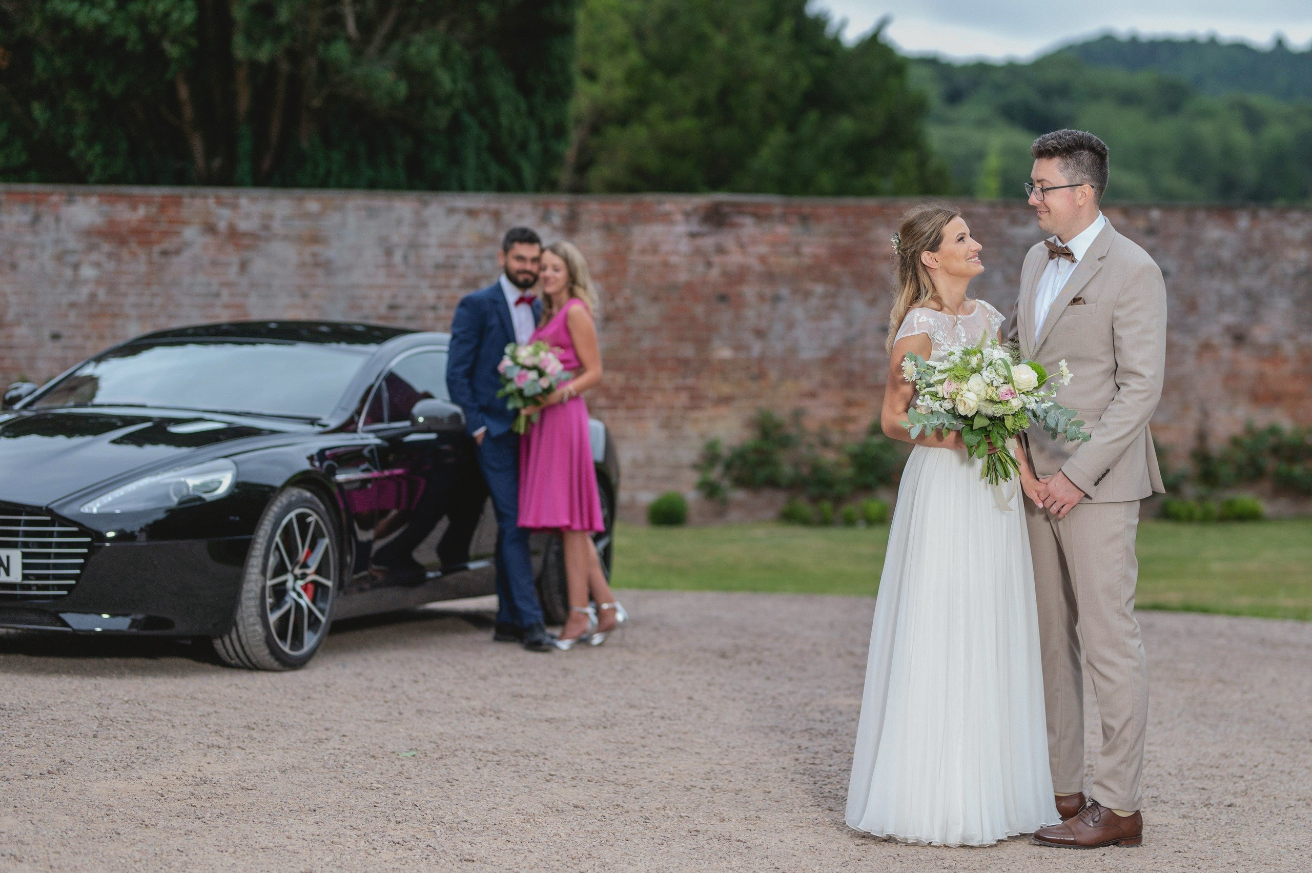 Bride and groom by the Aston Martin, with a bridesmaid and groomsman sharing a kiss in the background, Charlecote Pheasant Hotel