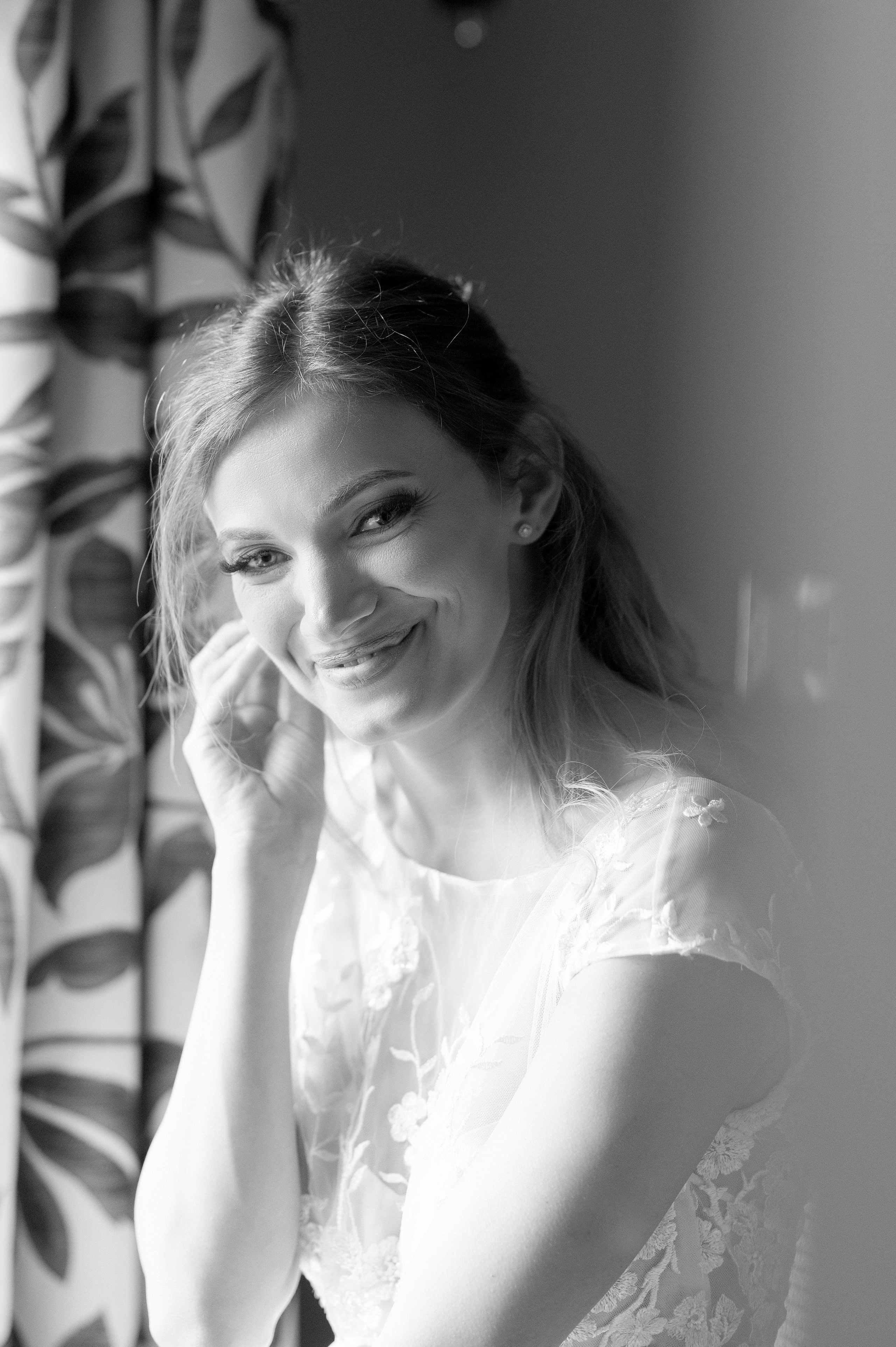 Bride smiling and fastening her earring by the window before the ceremony (black and white)