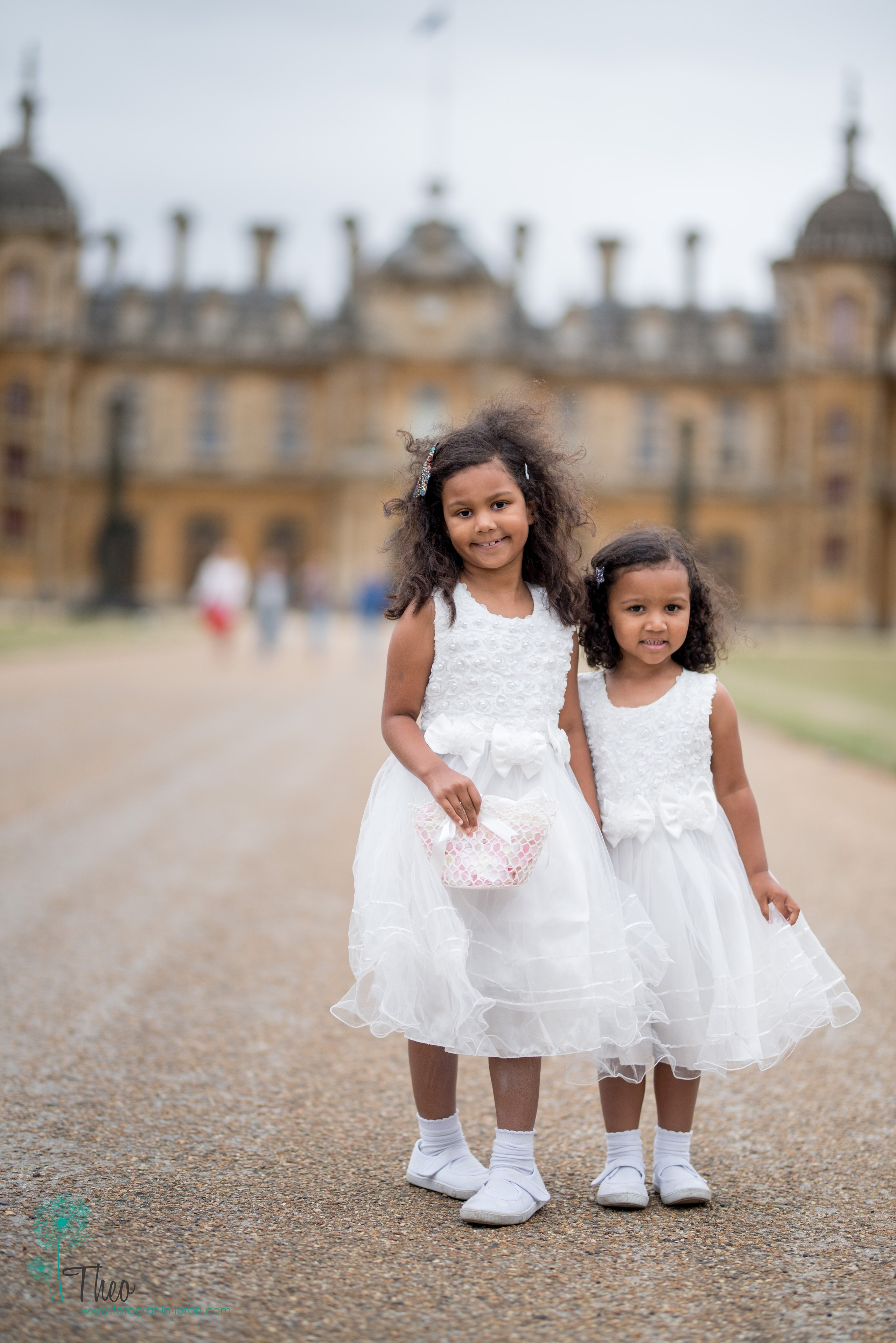 Two flower girls in white dresses, one carrying a flower basket, pose on the drive with Waddesdon Manor's towers behind them and visitors strolling past