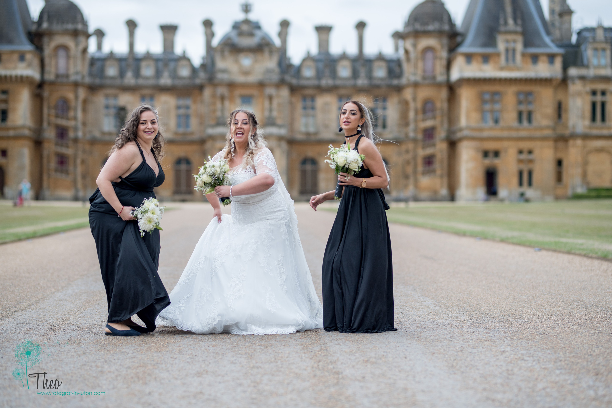 Bride laughs with her two bridesmaids in black gowns on the gravel drive at Waddesdon Manor, Buckinghamshire