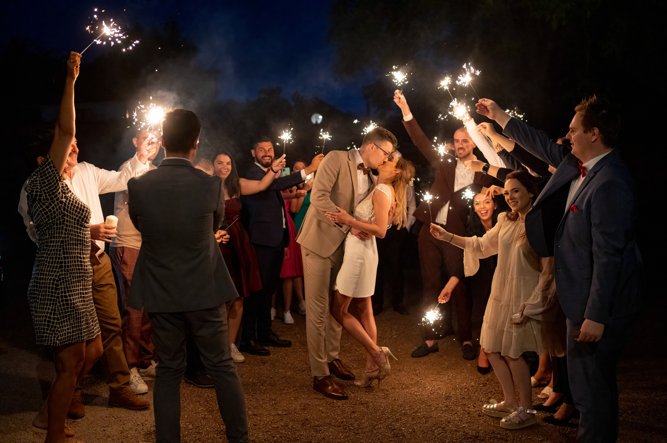 Bride and groom kissing in a sparkler tunnel surrounded by cheering guests at night