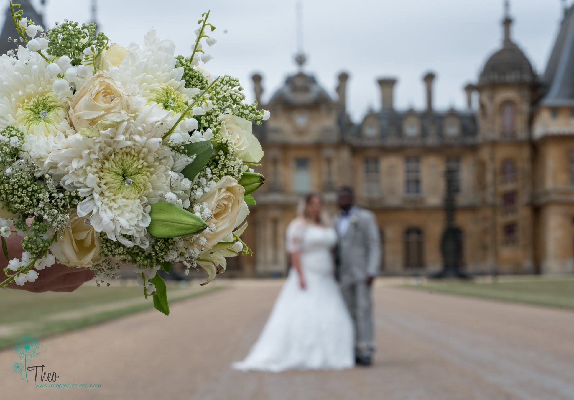 The bride's bouquet of cream roses and white chrysanthemums in sharp focus, with the newlyweds blurred in front of Waddesdon Manor