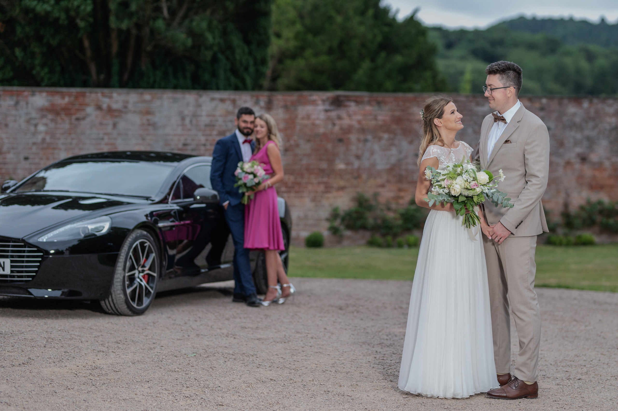 Bride and groom by the Aston Martin, with a bridesmaid and groomsman sharing a kiss in the background