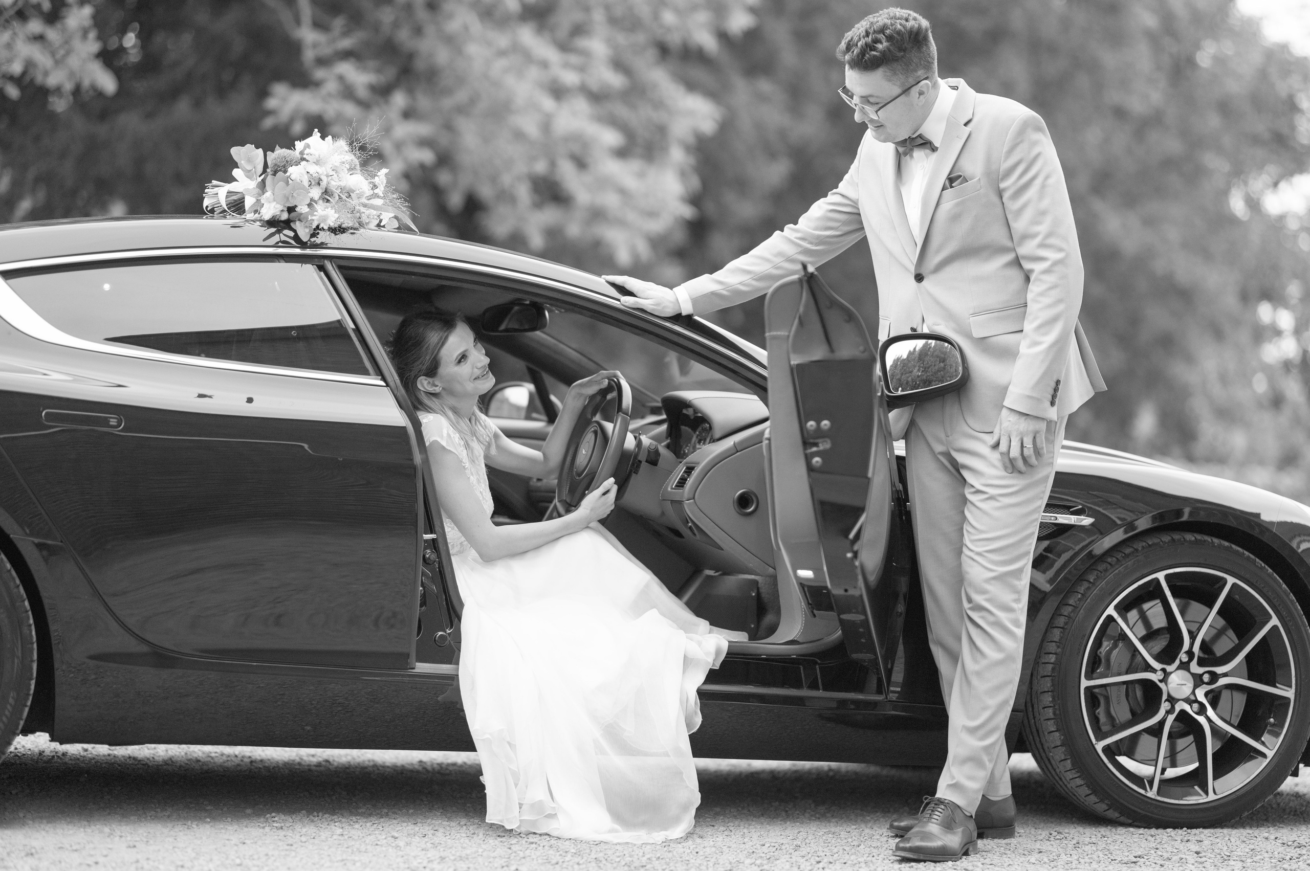 Groom laughing with the bride seated in the Aston Martin wedding car, bouquet on the roof (black and white)