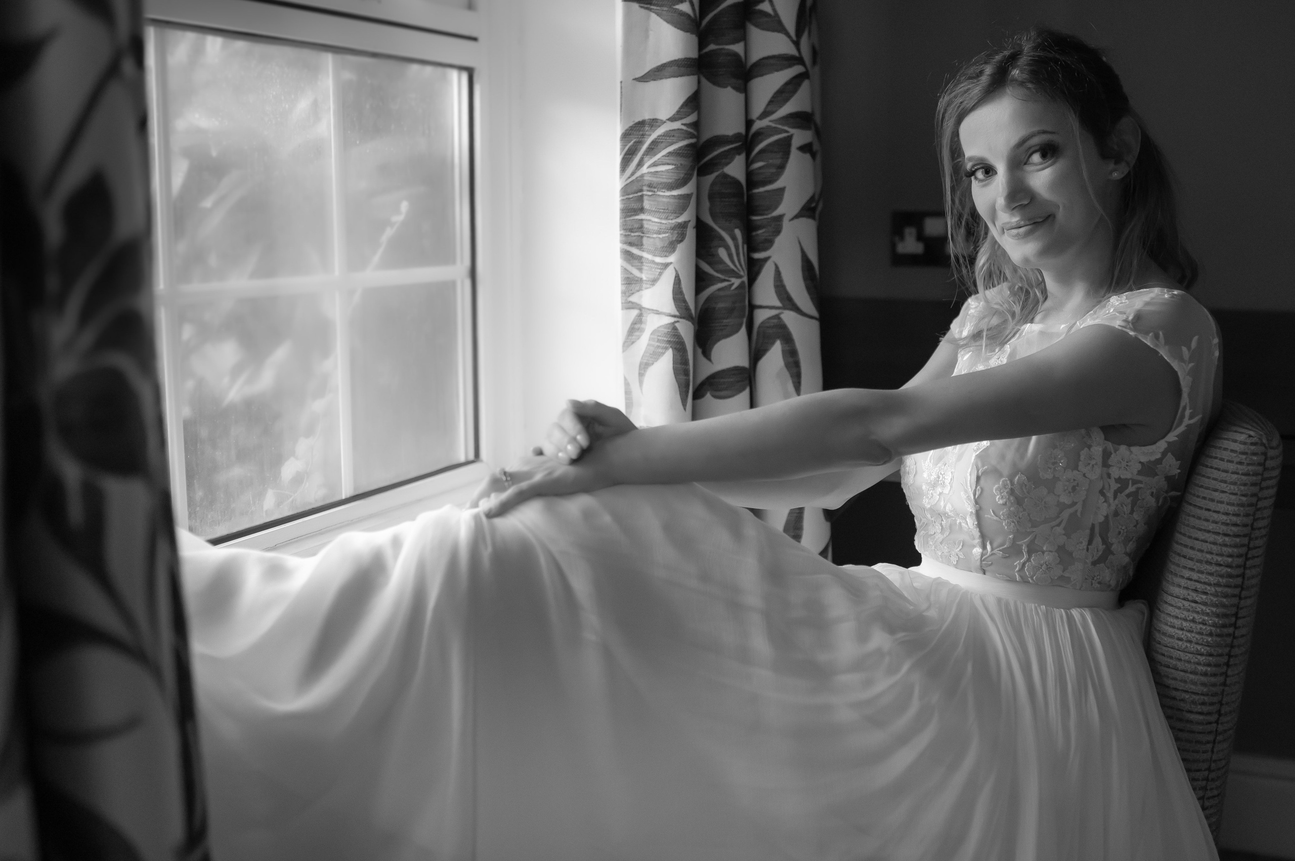 Bride relaxing by the window in her dress, smiling over her shoulder (black and white)