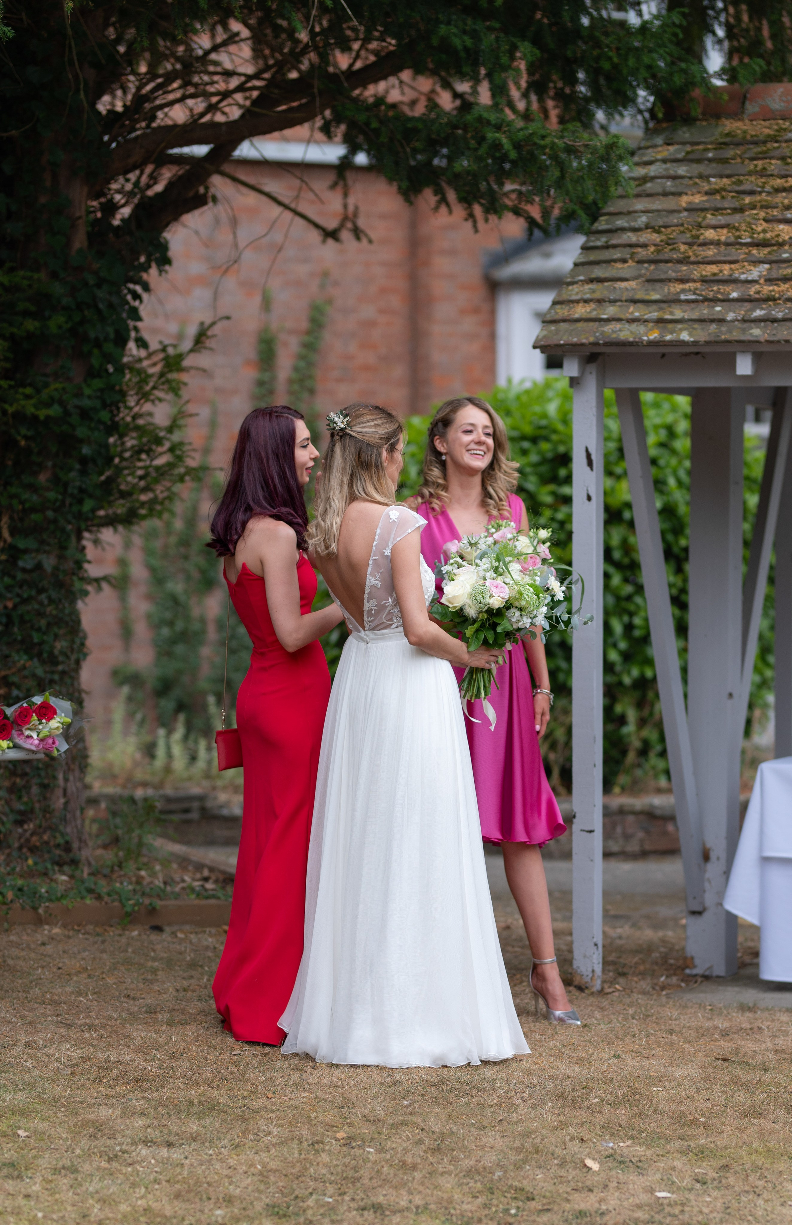 Bride chatting with bridesmaids in red and pink dresses by the garden gazebo after the ceremony