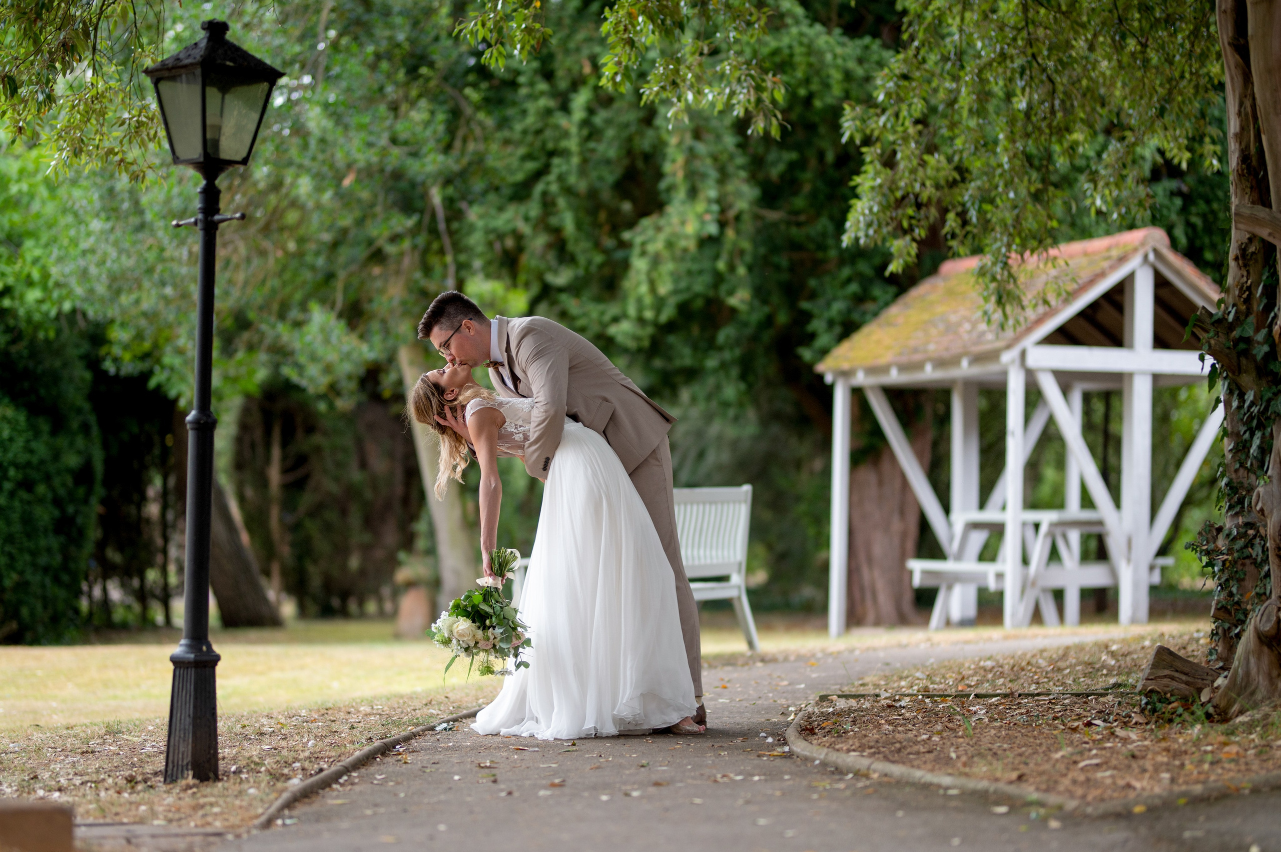 Wide shot of the romantic dip kiss under the trees, lamp post and garden shelter in the background