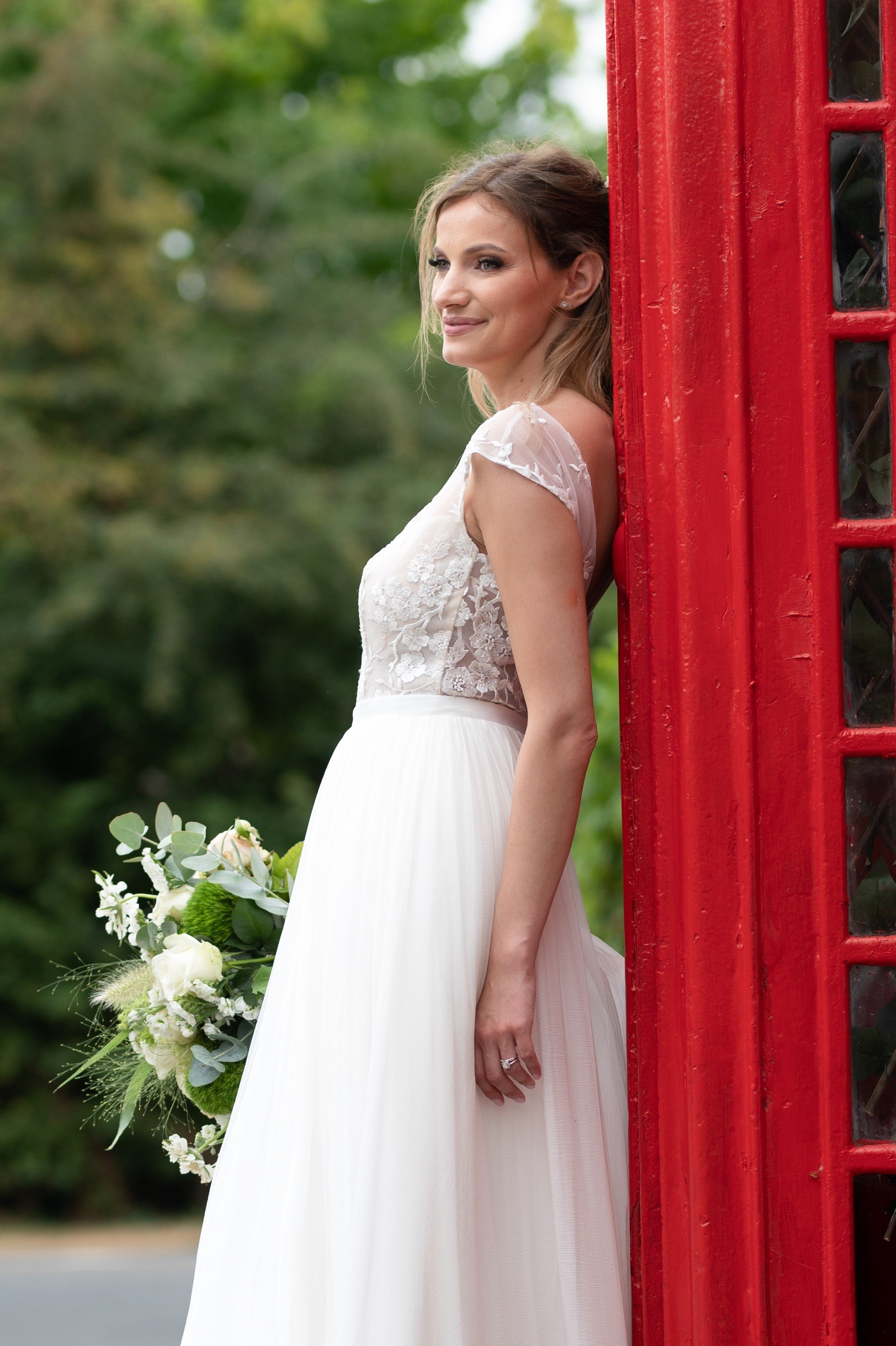 Bride leaning against a classic red British phone box, holding her bouquet, smiling into the distance