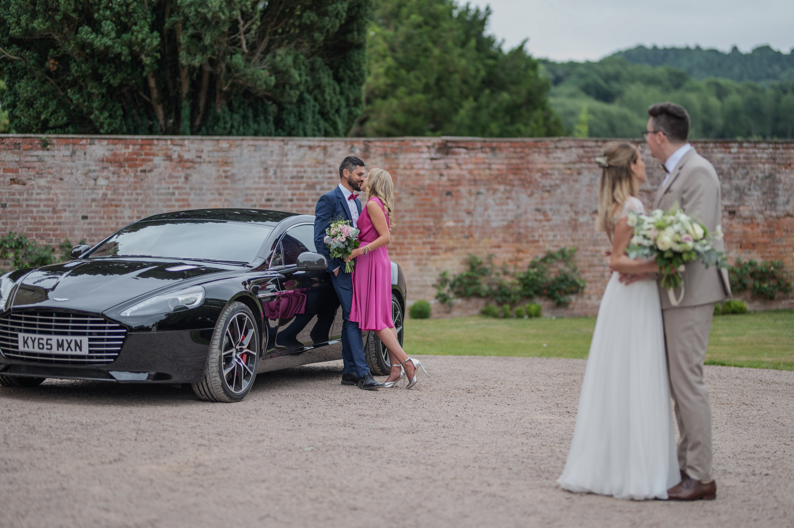 Bridesmaid and groomsman kissing by the wedding car, with the bride and groom looking on