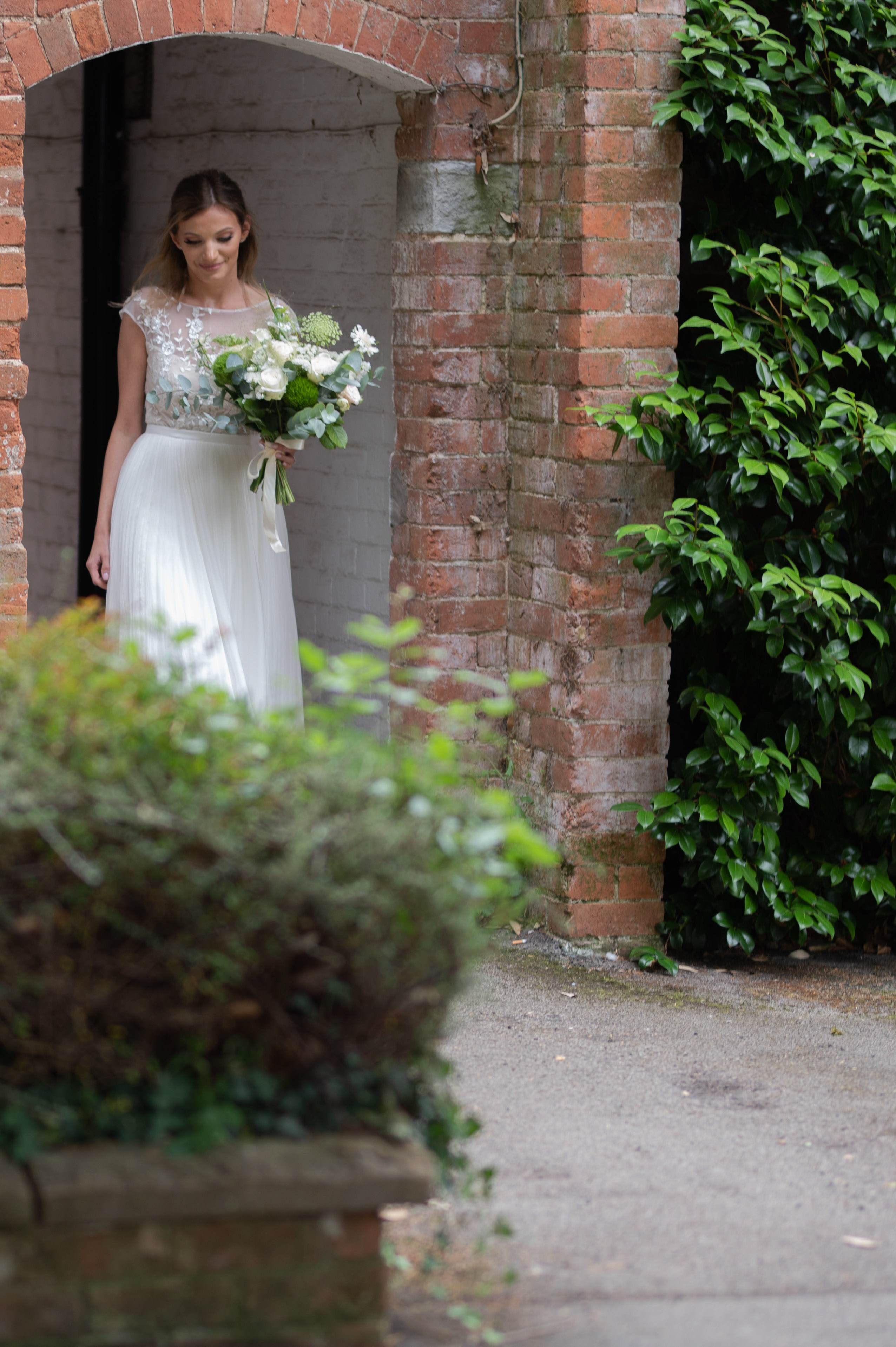 Bride in a white dress with a green and white bouquet, walking through a brick archway at Charlecote Pheasant Hotel