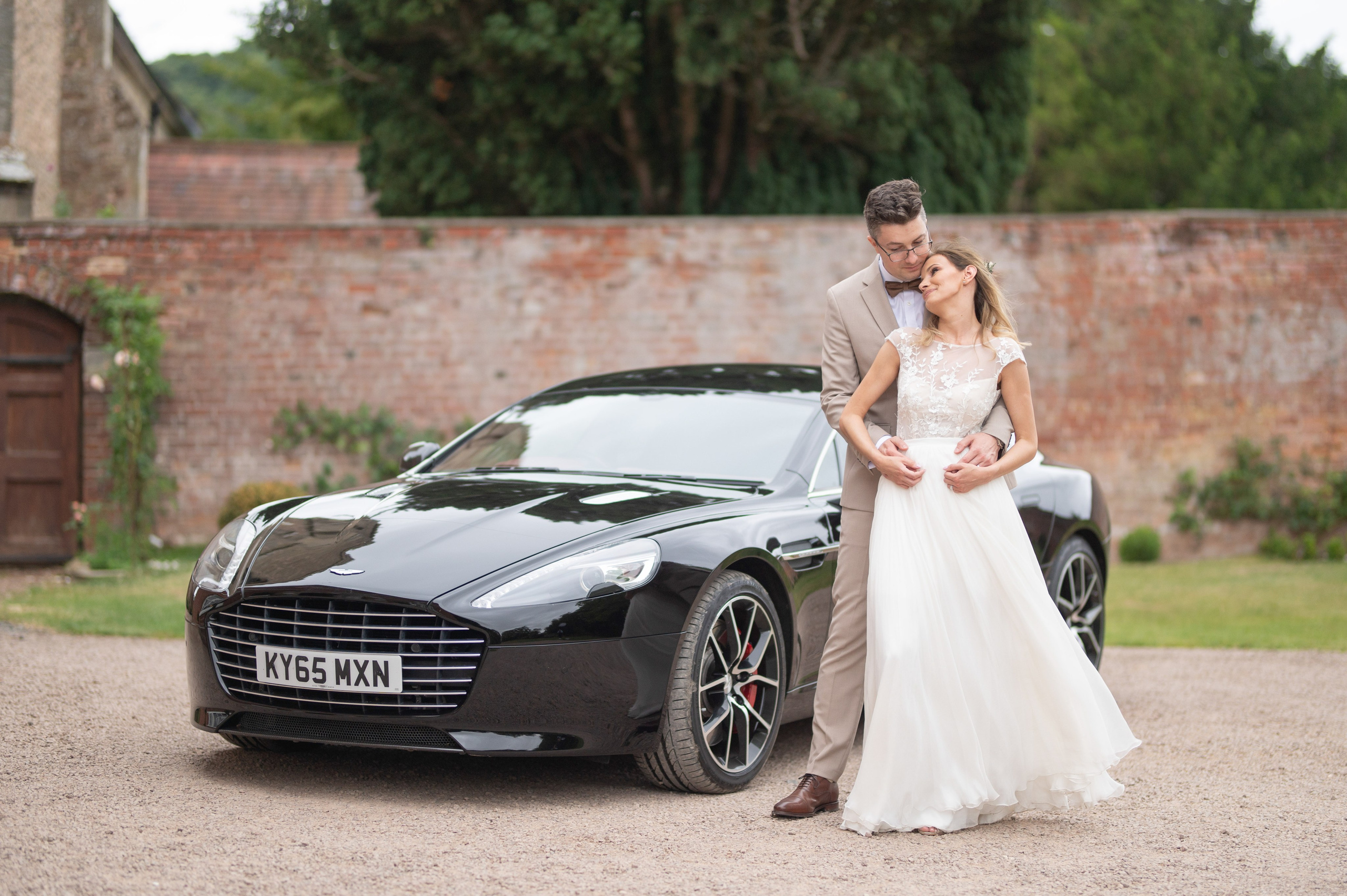 Bride and groom embracing beside their black Aston Martin wedding car