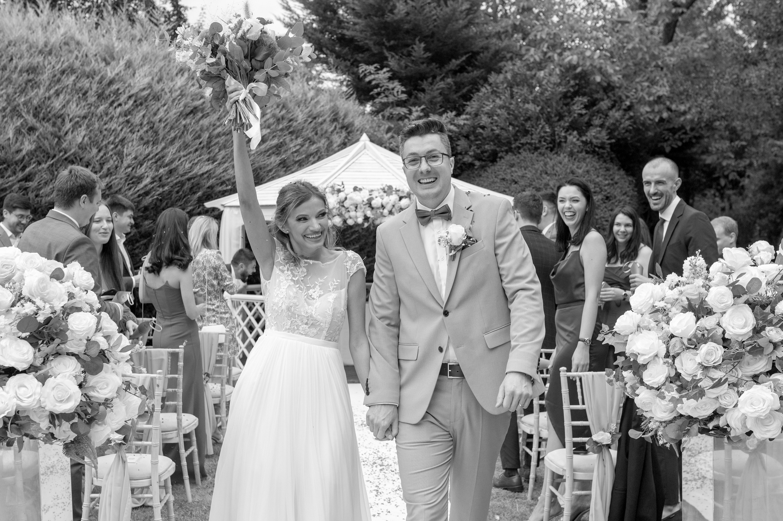 Newlyweds walking back down the aisle, bride raising her bouquet in celebration as guests cheer (black and white)
