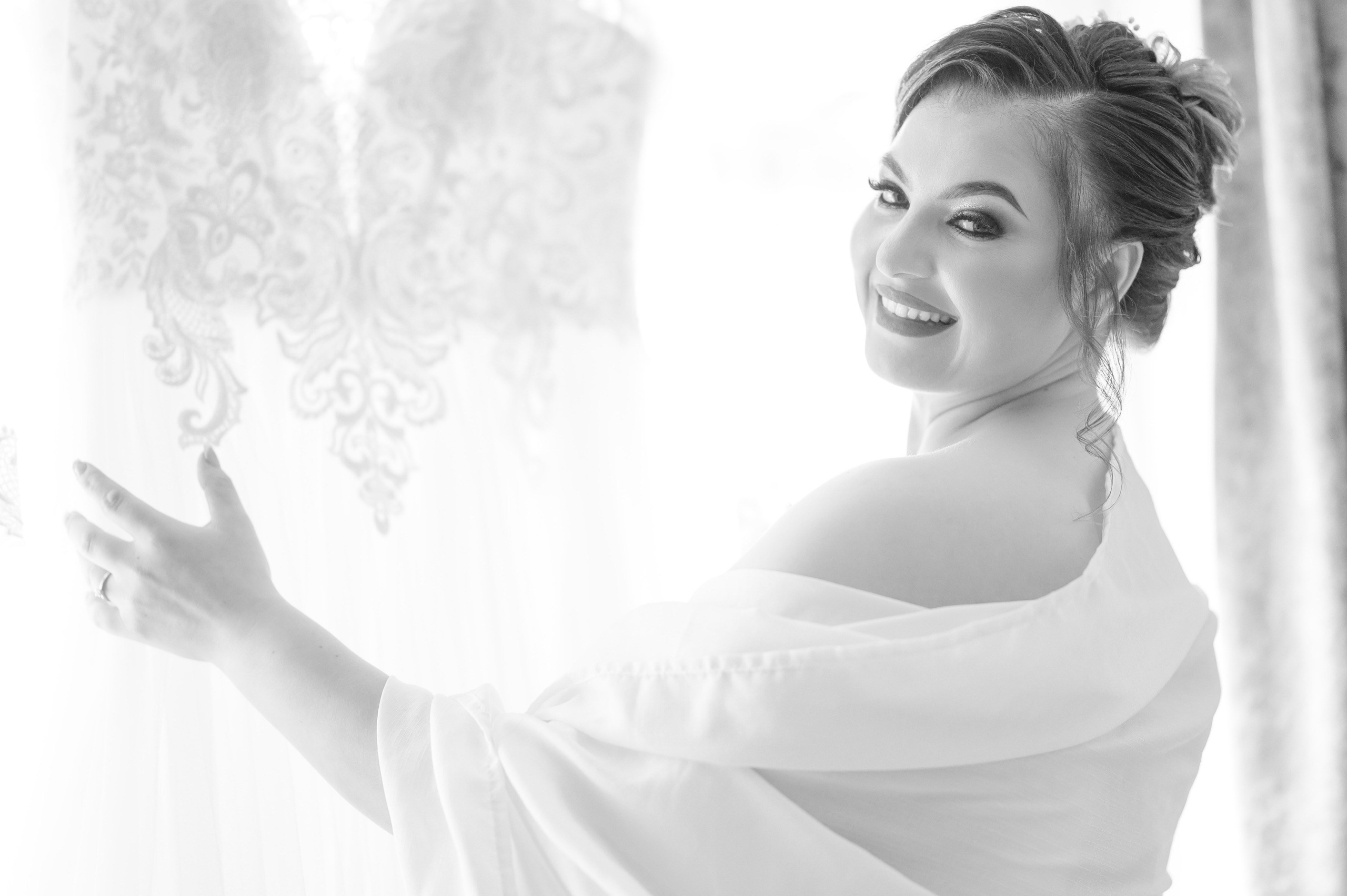 Bride in a lace wedding dress glancing back over her shoulder beside a sheer curtain, black and white portrait